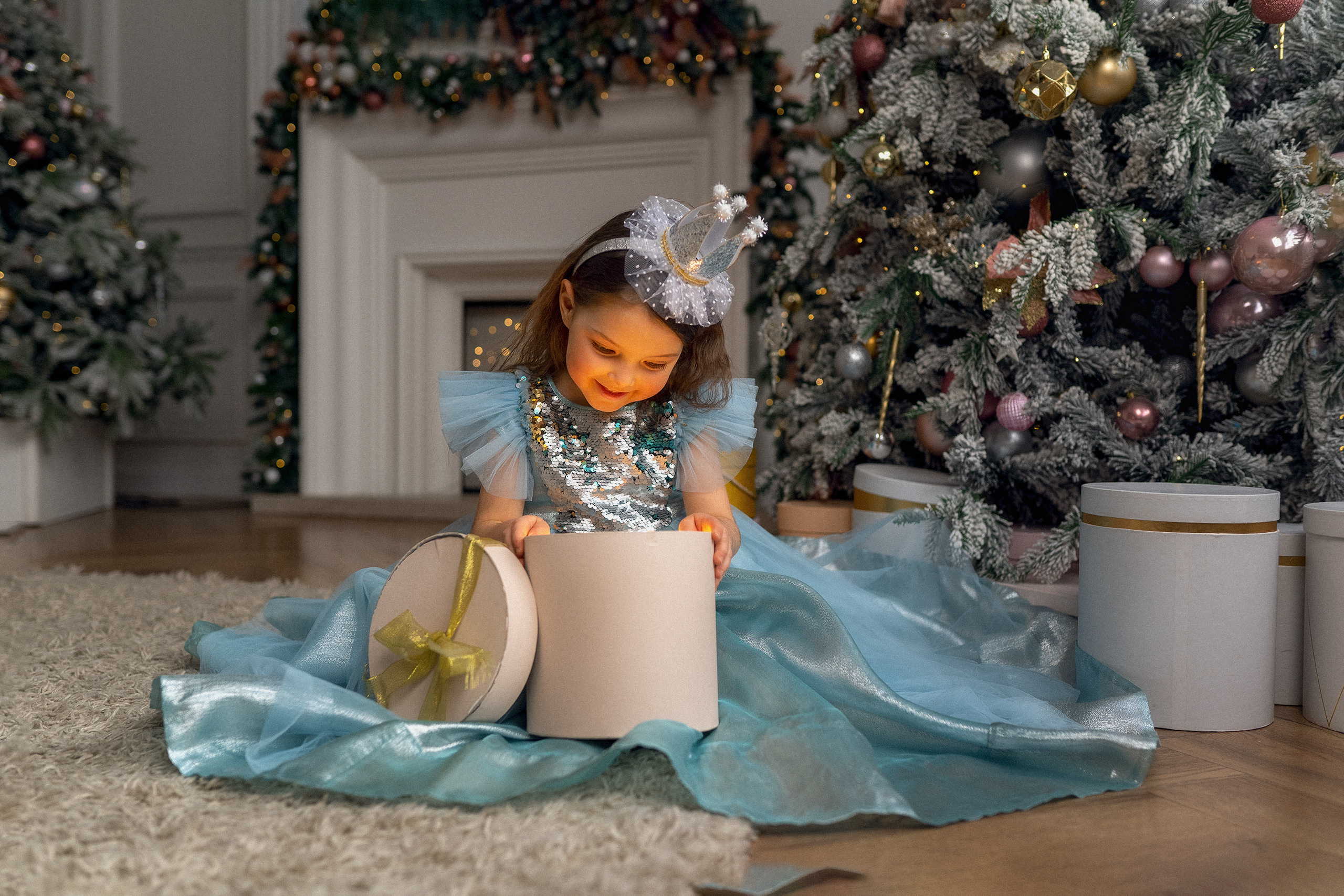 Маленькая девочка в платье принцессы с короной на голове открывает новогодний подарок на фоне камина новогодней елки и подарочных коробок