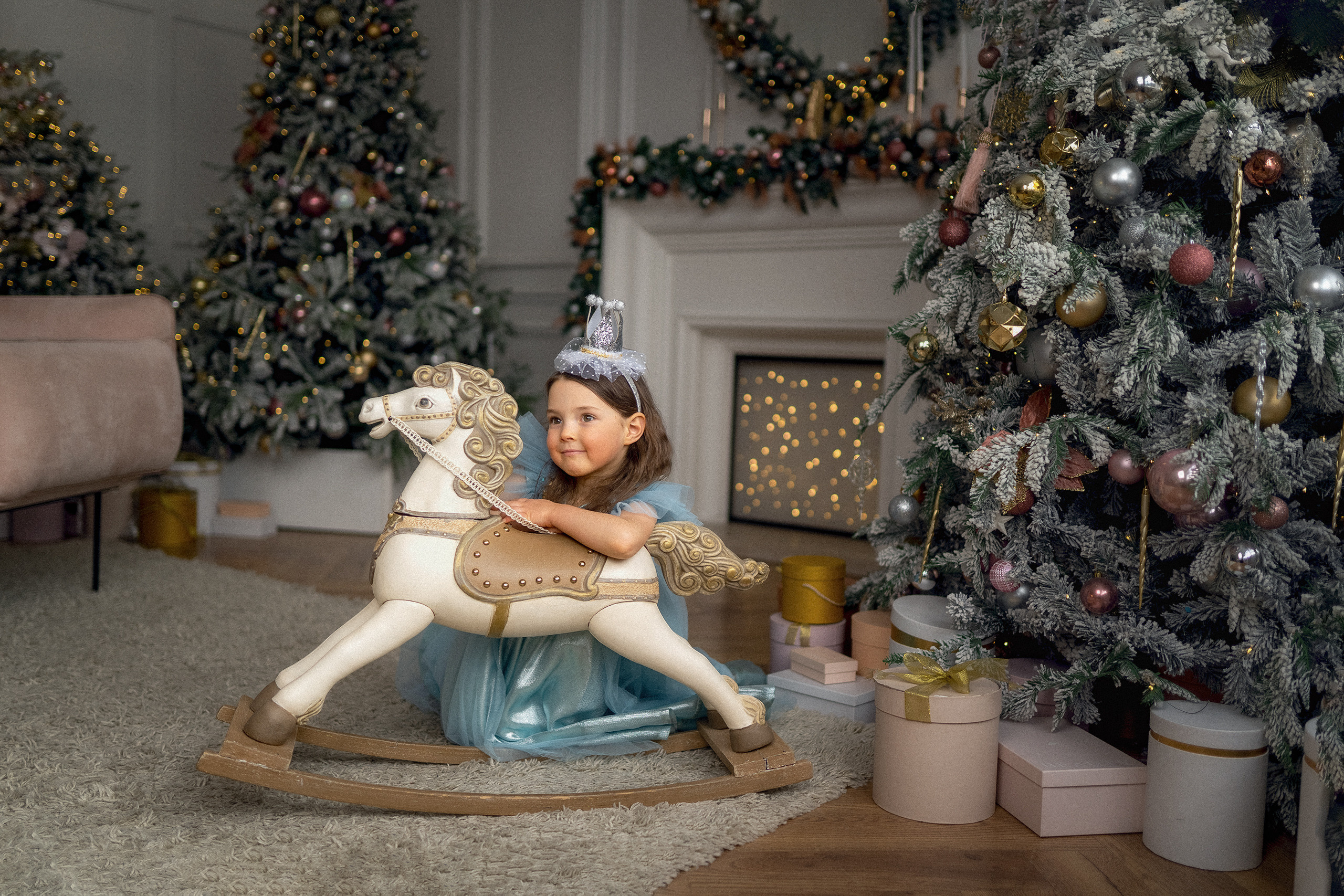 Маленькая девочка в платье принцессы с короной на голове с деревянной лошадкой качалкой на фоне камина новогодней елки и подарочных коробок