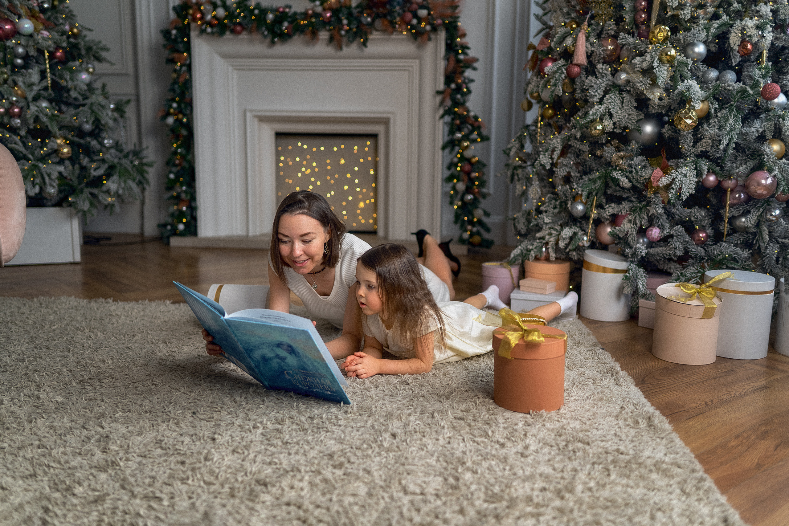 Мама с дочкой лежат на ковре и читают книгу на фоне камина новогодней елки и подарочных коробок