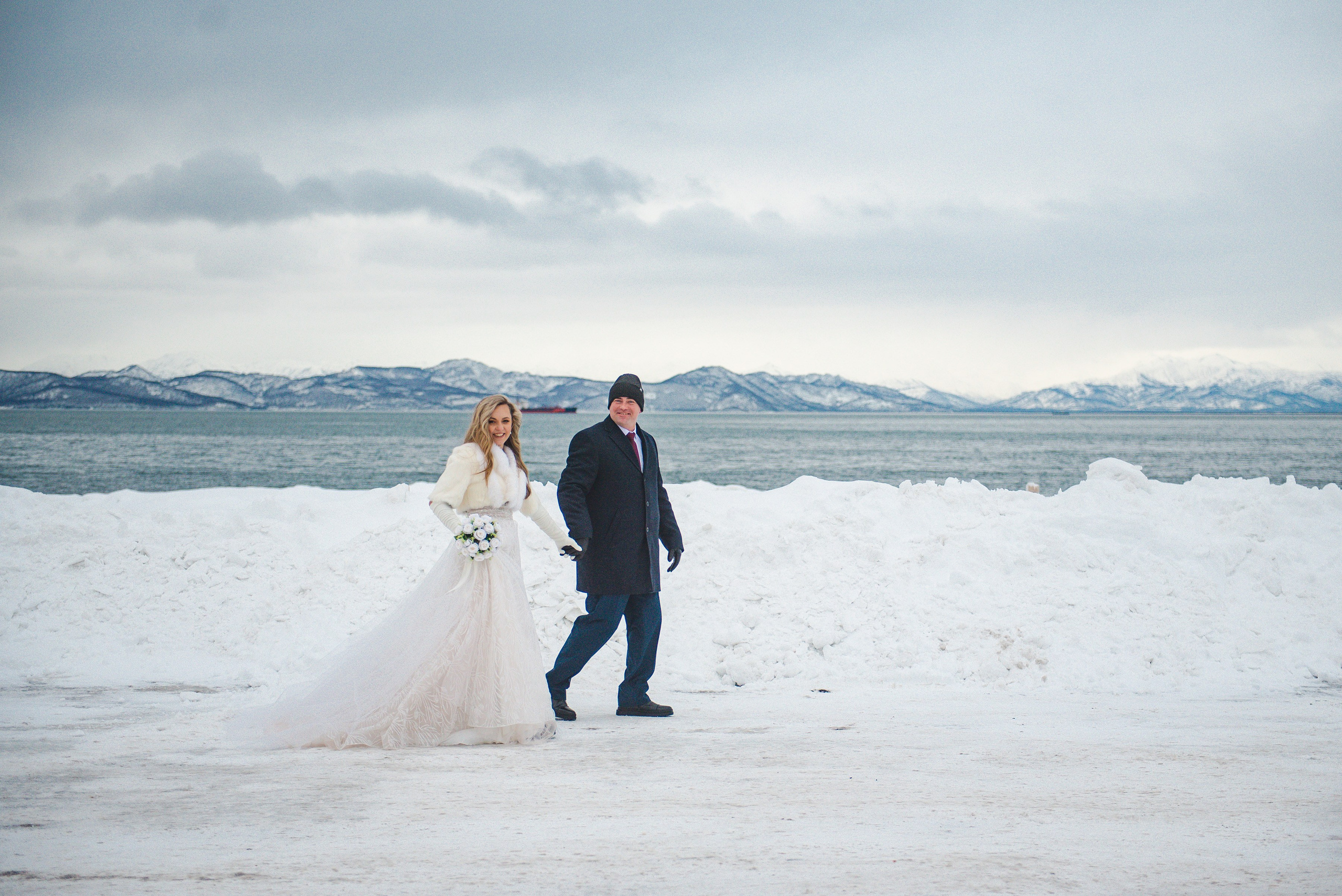 Невеста в пышном белом платье с букетом и жених в тёмном пальто идут по заснеженному берегу. На фоне — горы, море, пасмурное небо. Атмосфера зимняя, романтичная.
