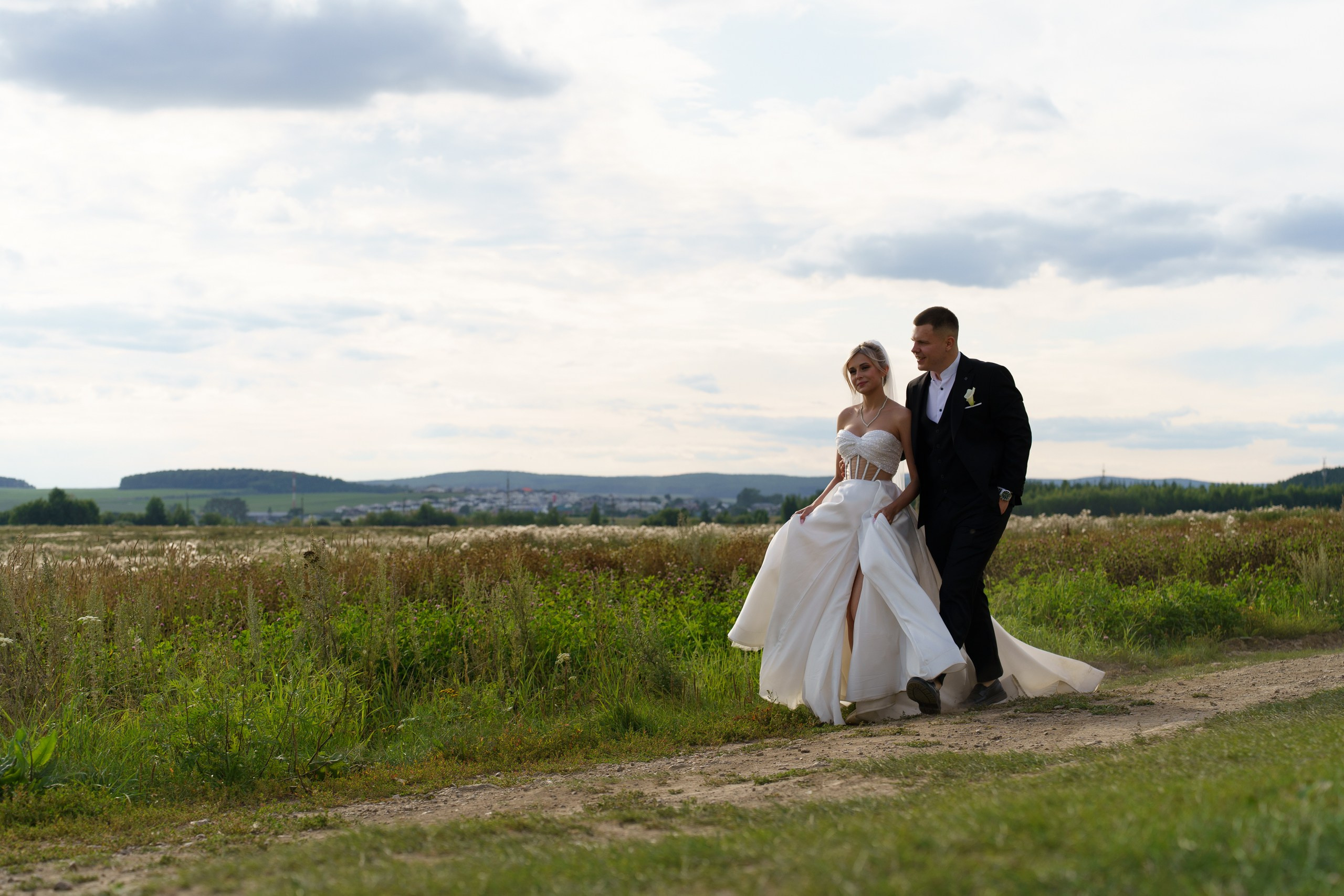 Жених в чёрном костюме и невеста в белом платье прогуливаются по просёлочной дороге. На заднем плане — зелёные поля, холмы и облачное небо, создающие романтическую атмосферу | вечерняя свадебная фотосессия Екатеринбург