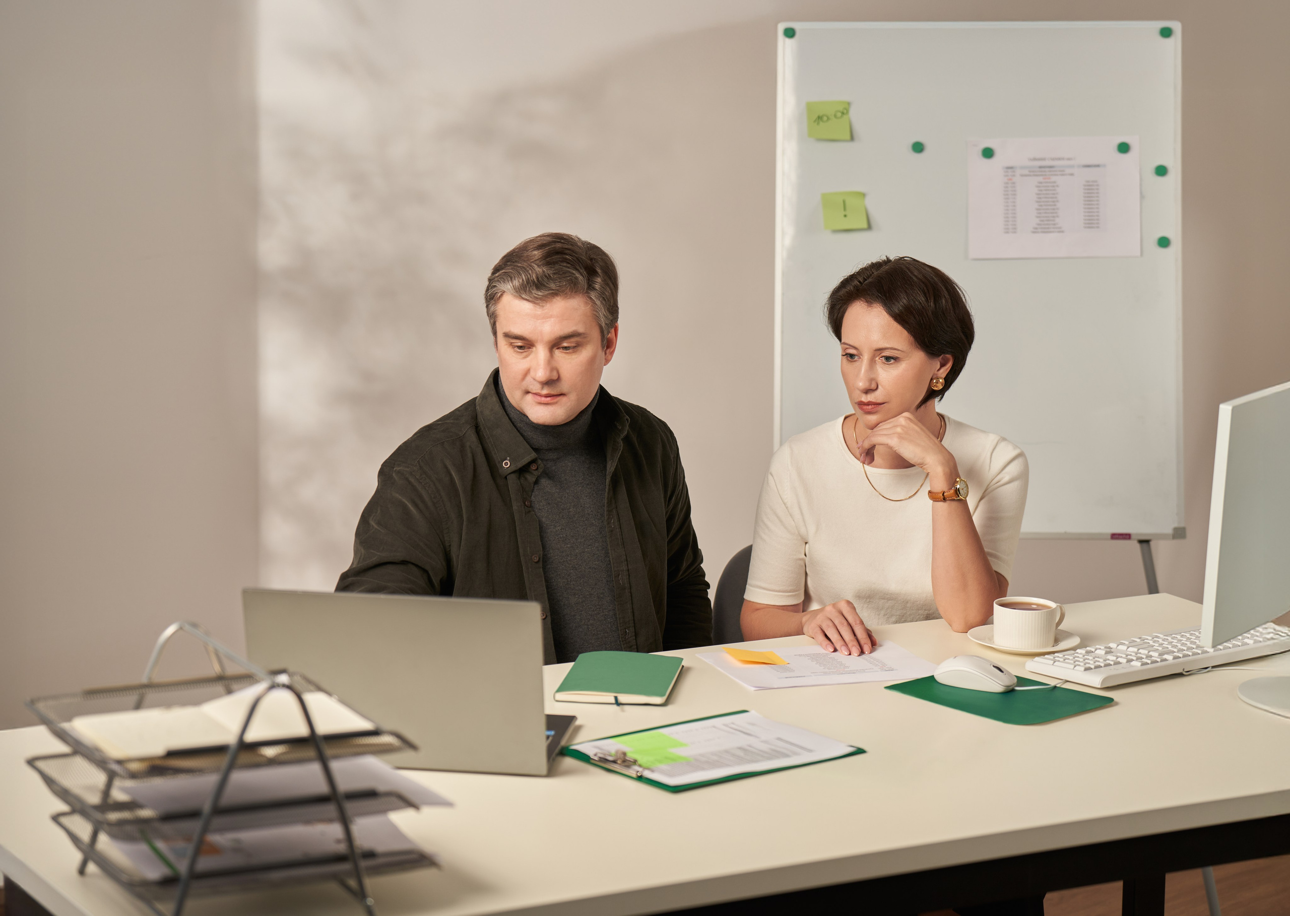 Мужчина и женщина вместе работают за ноутбуком в офисе, на фоне доска с заметками