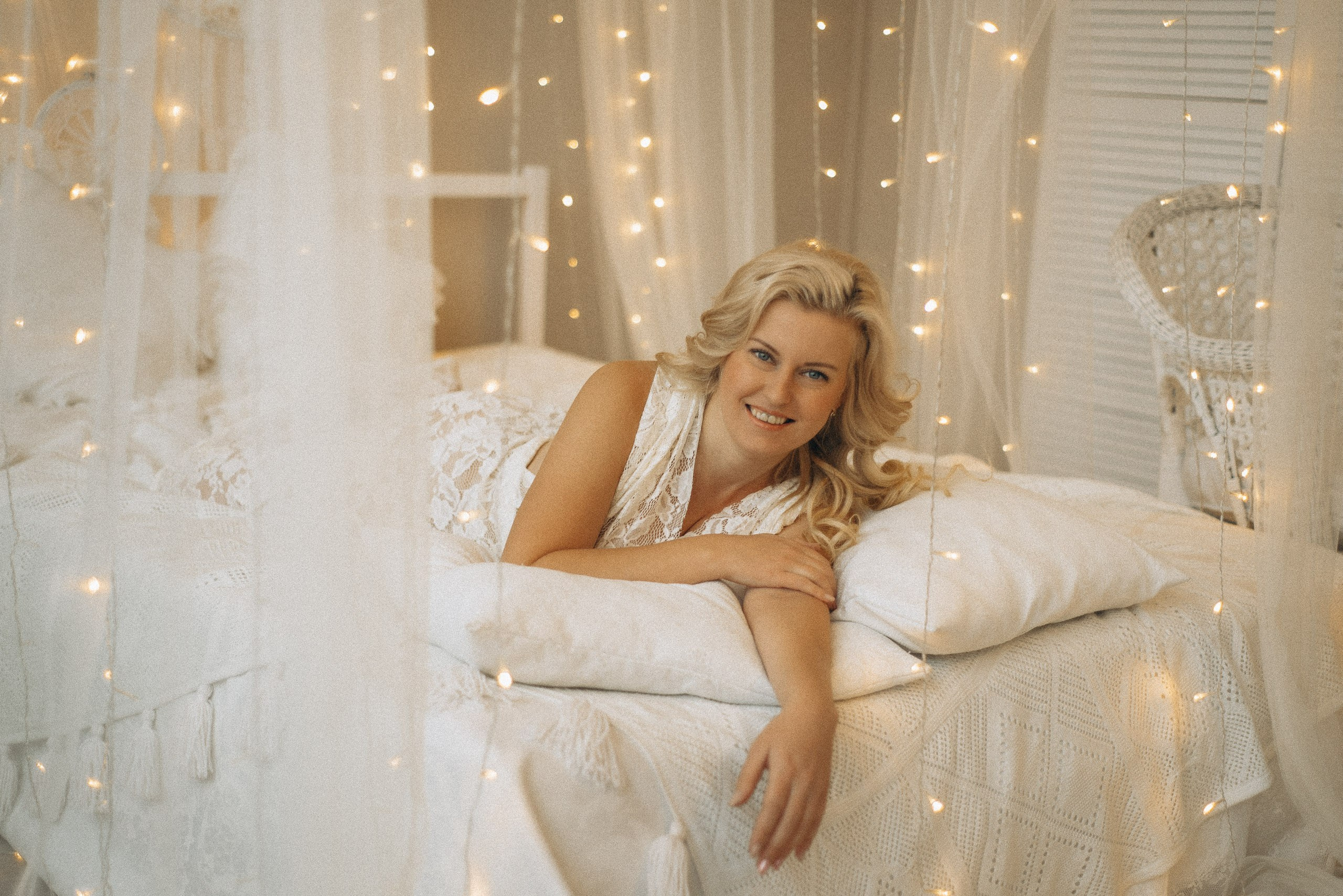 Улыбающаяся Натали Каролин на кровати в окружении фатина и огней. Модель в белом кружевном платье в интерьере бохо-студии. Атмосферная женская фотосессия в Новосибирске