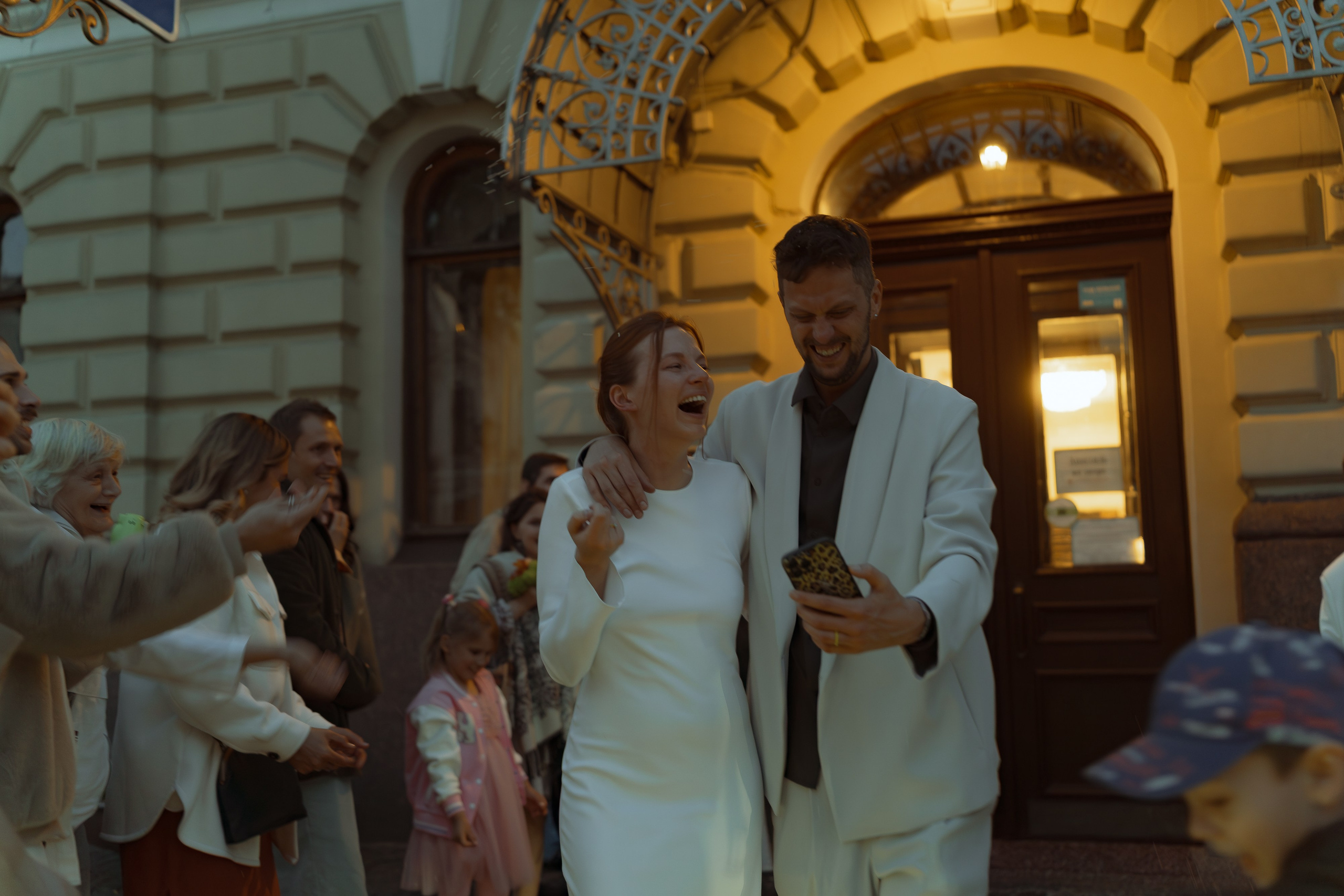 Мария смеётся, обняв Евгения, пока он показывает что-то на телефоне перед фасадом ЗАГСа