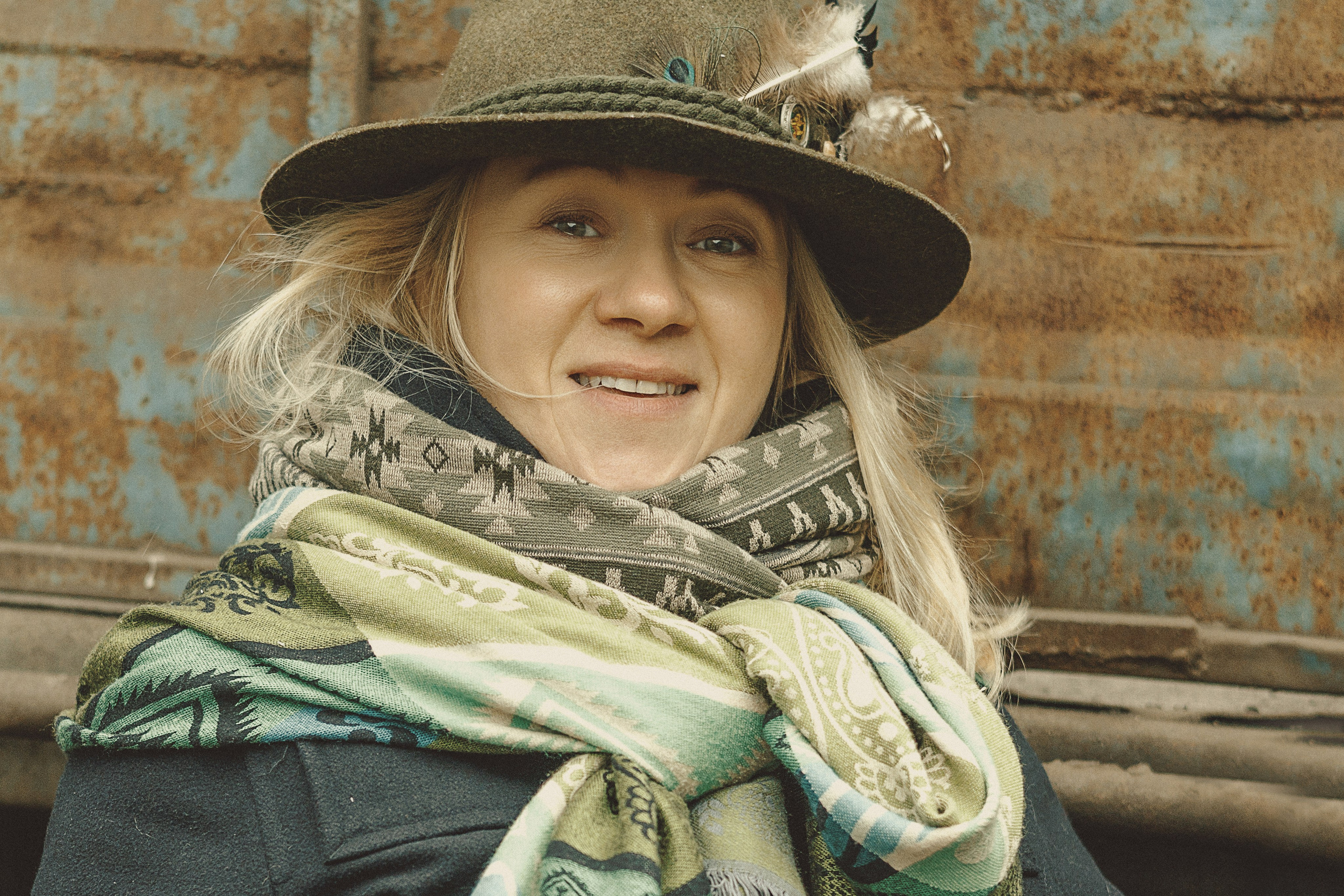 Женщина в шляпе с пером и узорчатом шарфе улыбается на улице на фоне ржавой металлической стены.