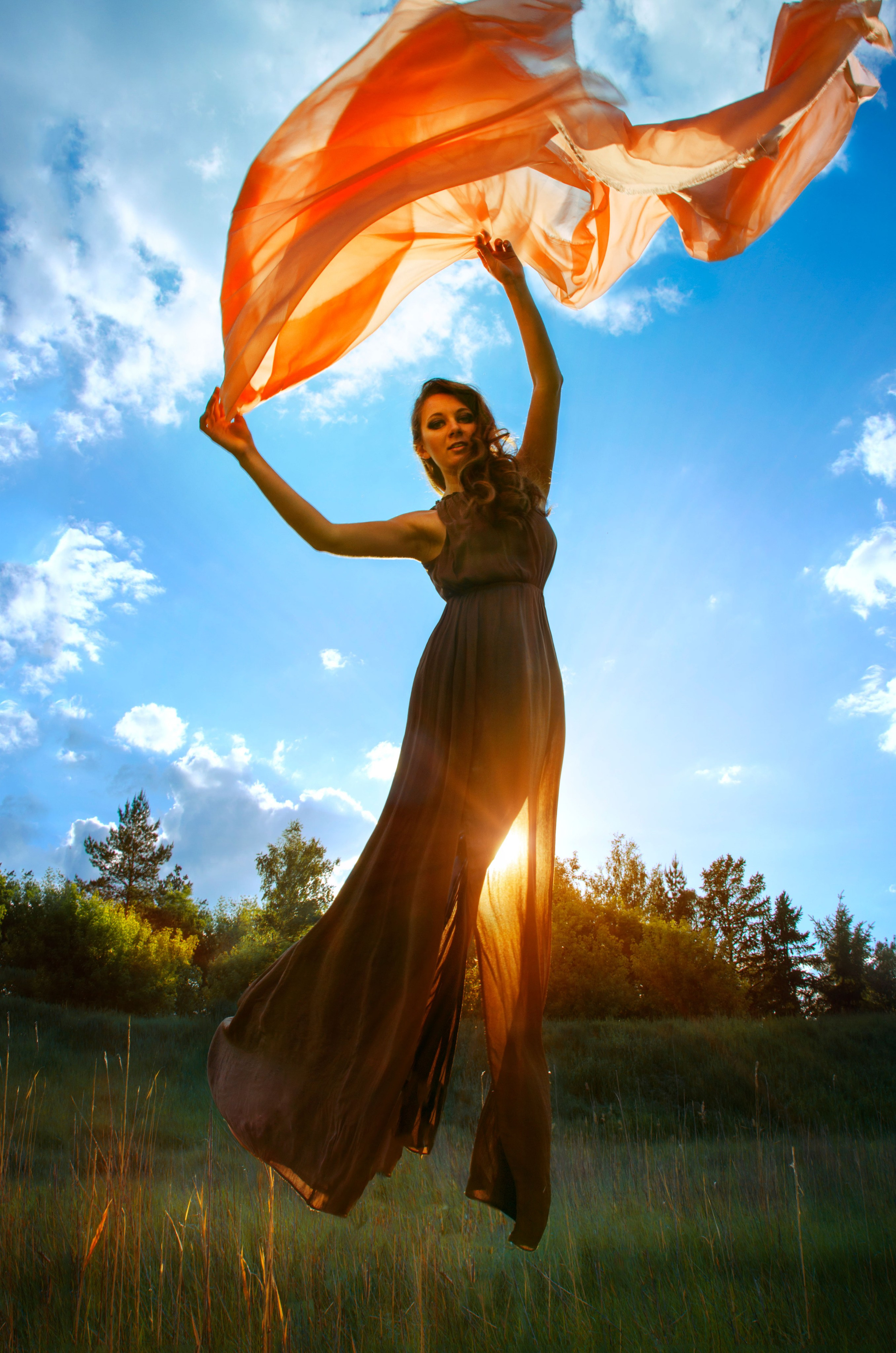 фотосессия с левитацией, девушка в платье летит на фоне голубого неба с оранжевой тканью в руках на фоне заката