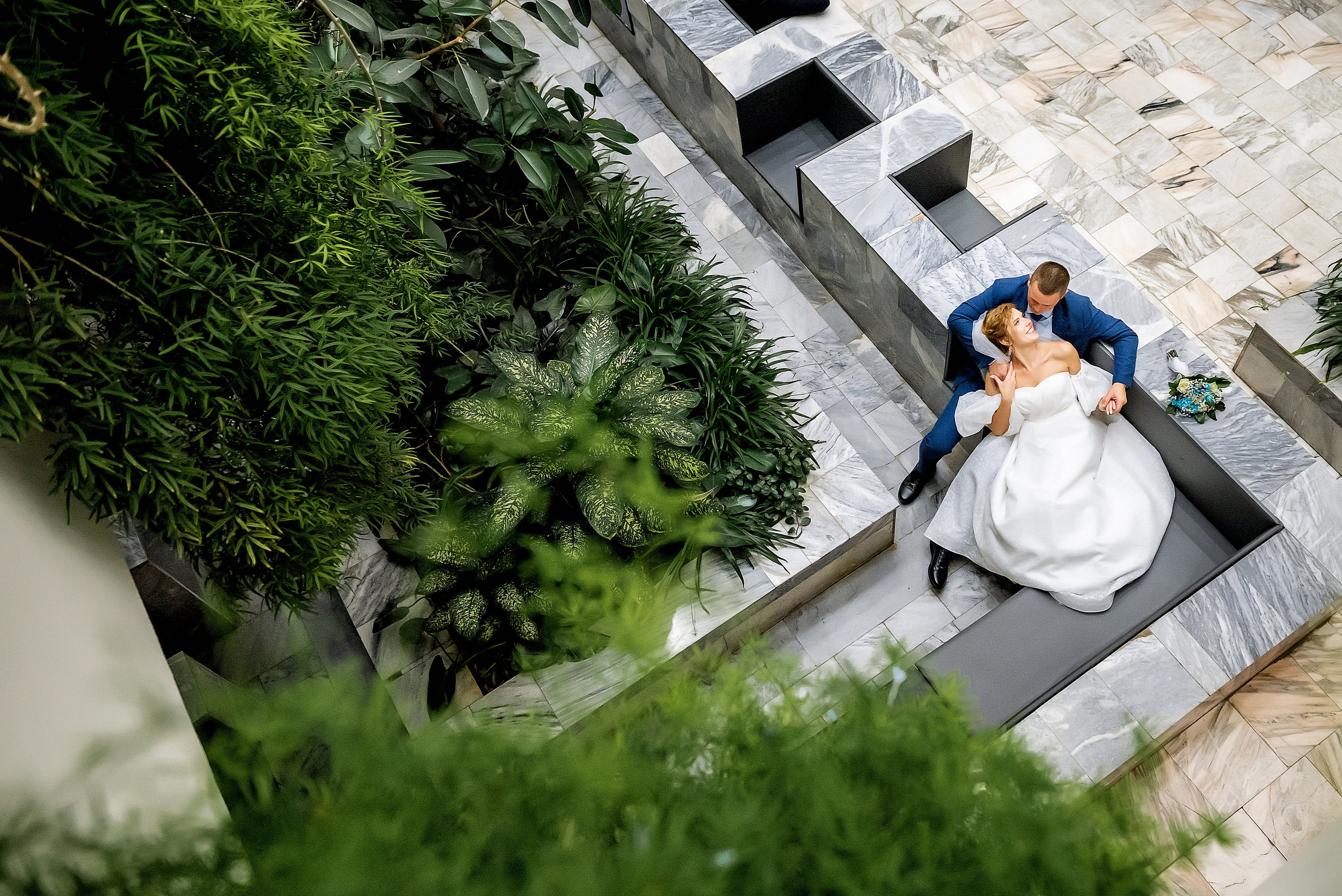 Свадебная пара сидит на современном декоративном диване в окружении зелени. Женщина в белом свадебном платье, а мужчина в синем костюме нежно держит её за руку, создавая романтичную атмосферу. Фон состоит из видов на зеленые растения и каменные поверхности. Снимок выполнен сверху, подчеркивая их близость и уют.
