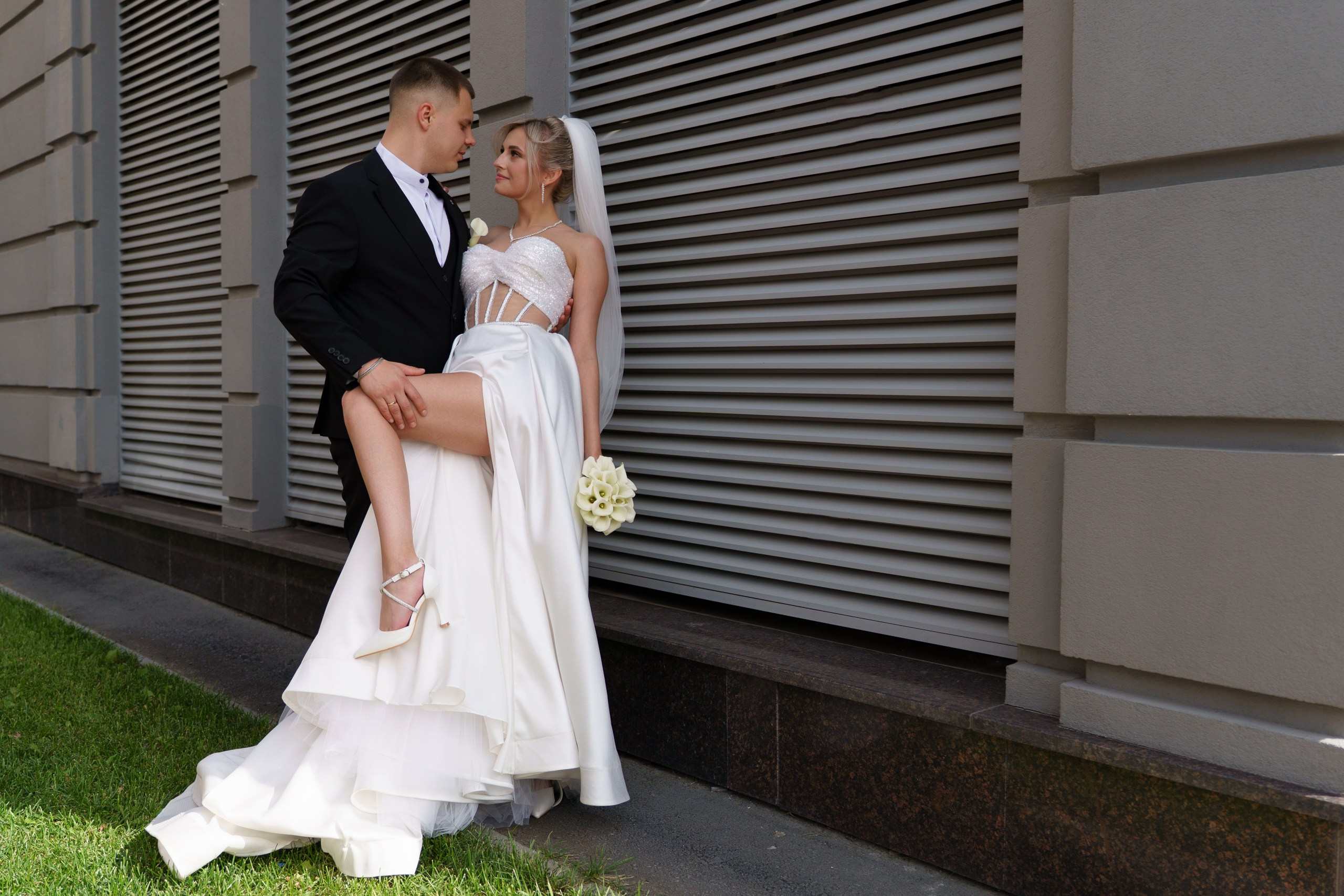 Свадебная фотосессия: невеста в роскошном белом платье с корсетным верхом и фатой и жених в классическом чёрном костюме запечатлены в романтичной и немного игривой позе. На заднем плане — современное здание | фотограф для запоминающейся свадьбы Екатеринбург