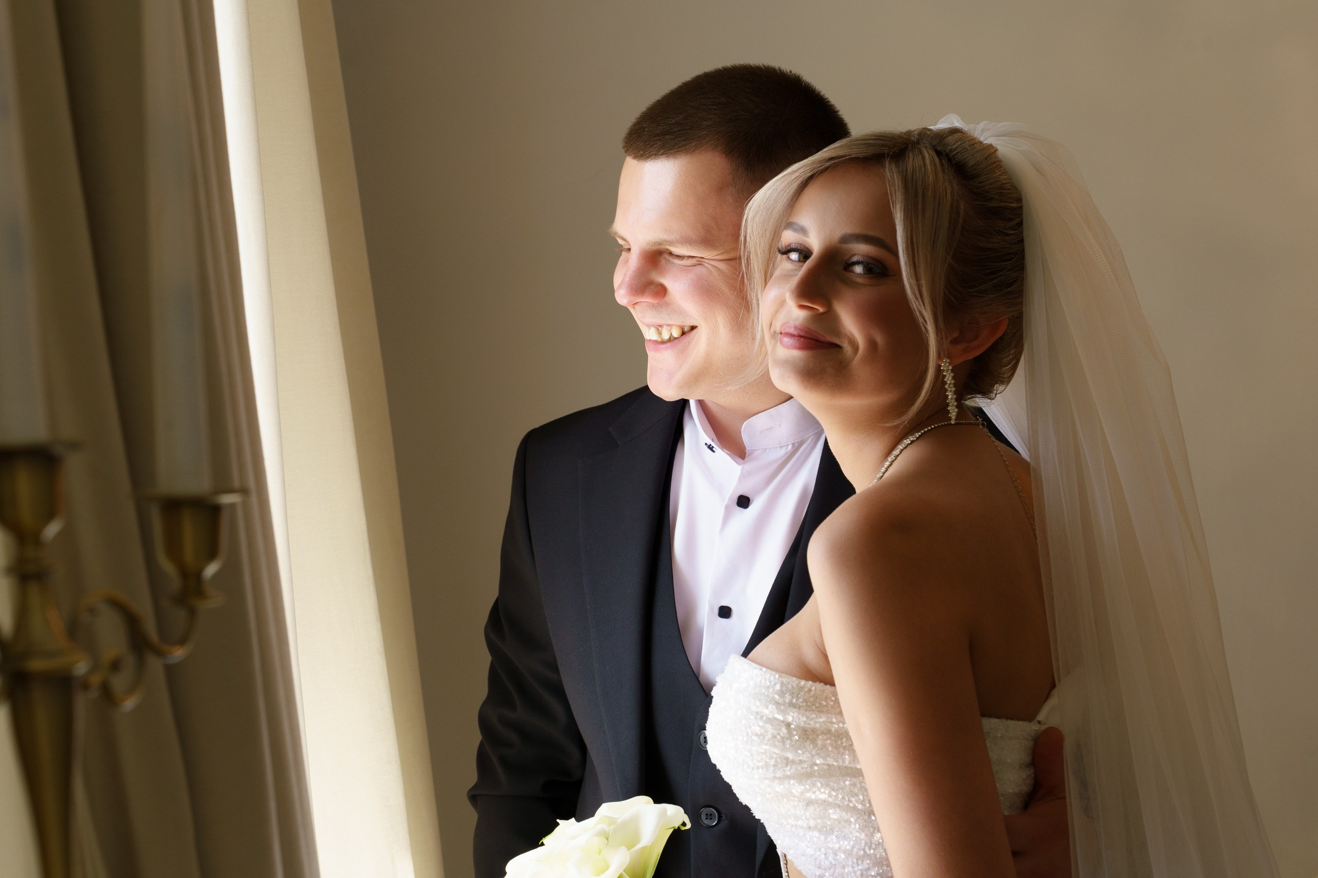 Свадебная фотосессия у окна: невеста в элегантном платье и фате держит букет белых роз, рядом с ней — улыбающийся жених в классическом костюме | фотограф для свадебного банкета Екатеринбург