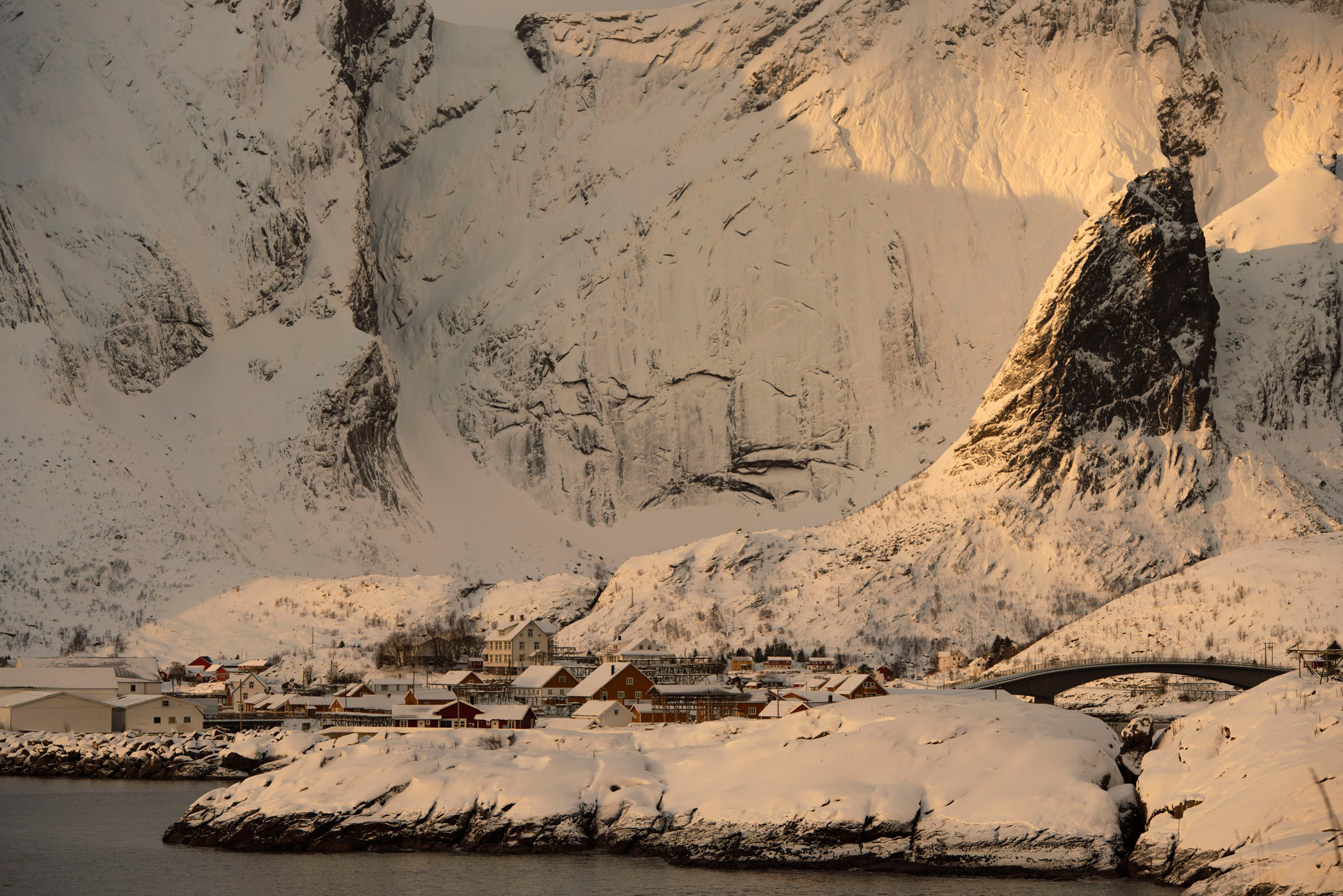 Зимний пейзаж норвежской деревни Рейне (Reine) на Лофотенских островах, расположенной у подножия величественных заснеженных гор, с мостом и водой на переднем плане.