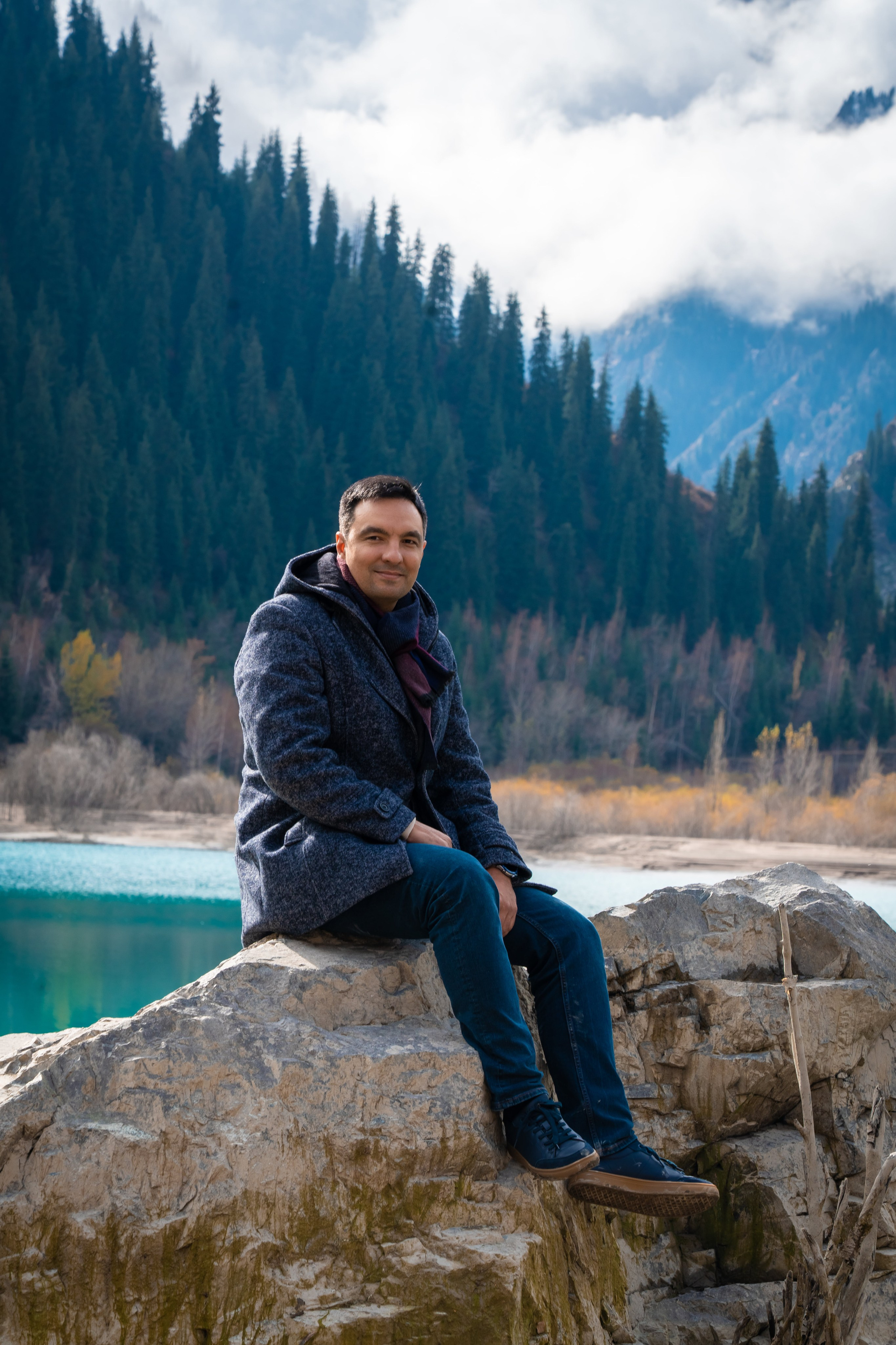 Путешественник компании Melon Travel сидит на большом камне на фоне красивого горного пейзажа. Позади него — бирюзовое озеро Иссык, густой хвойный лес и горы, частично скрытые облаками. Он одет в теплую одежду: серое пальто и шарф, создавая атмосферу осеннего или раннего зимнего дня.