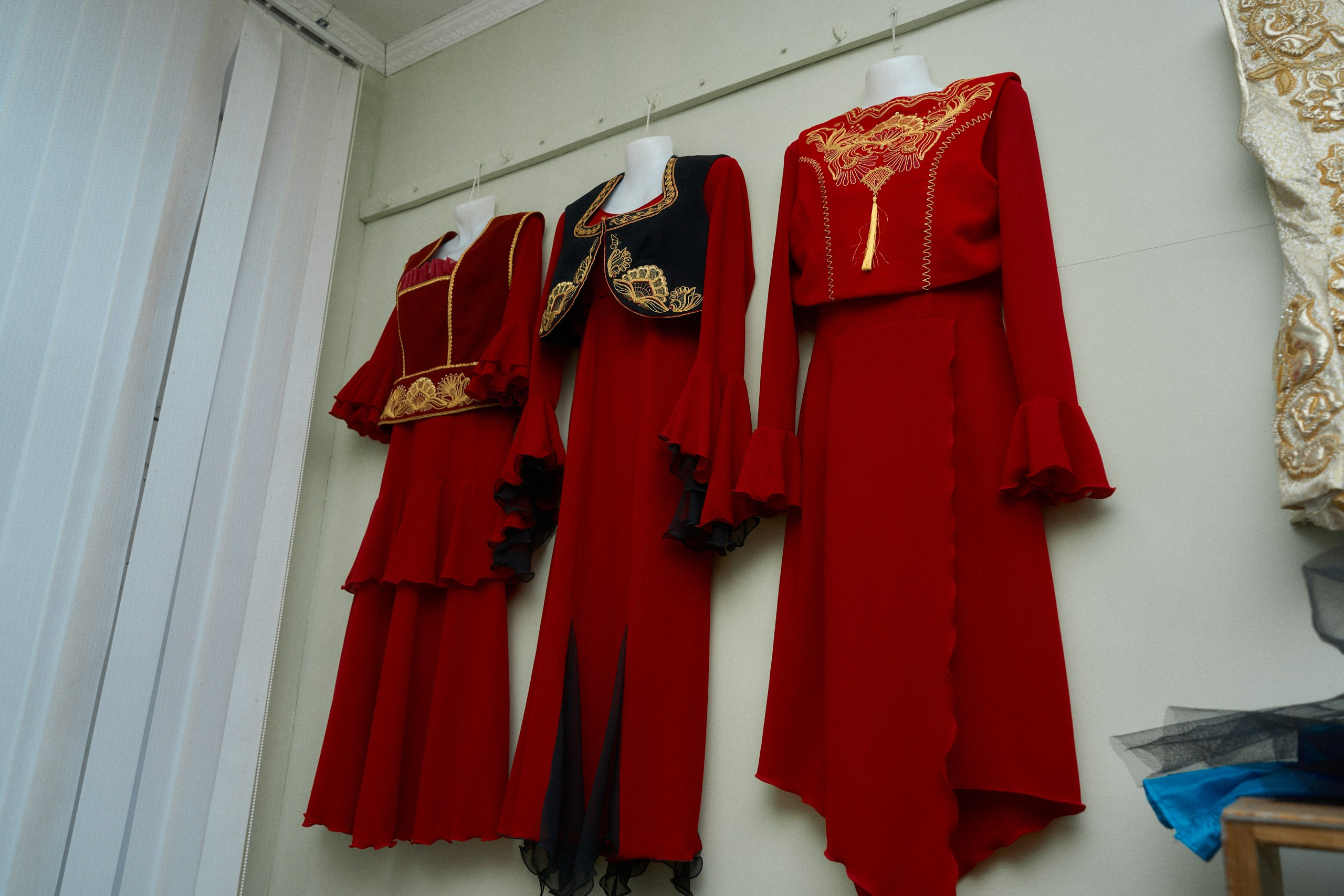 Три традиционных женских костюма, выставленных на стене в музее. Костюмы выполнены в ярко-красных тонах с богатой золотой вышивкой на воротниках, рукавах и подолах. Центральный костюм имеет черные вставки, украшенные золотыми узорами, что подчеркивает контраст и изысканность вышивки.