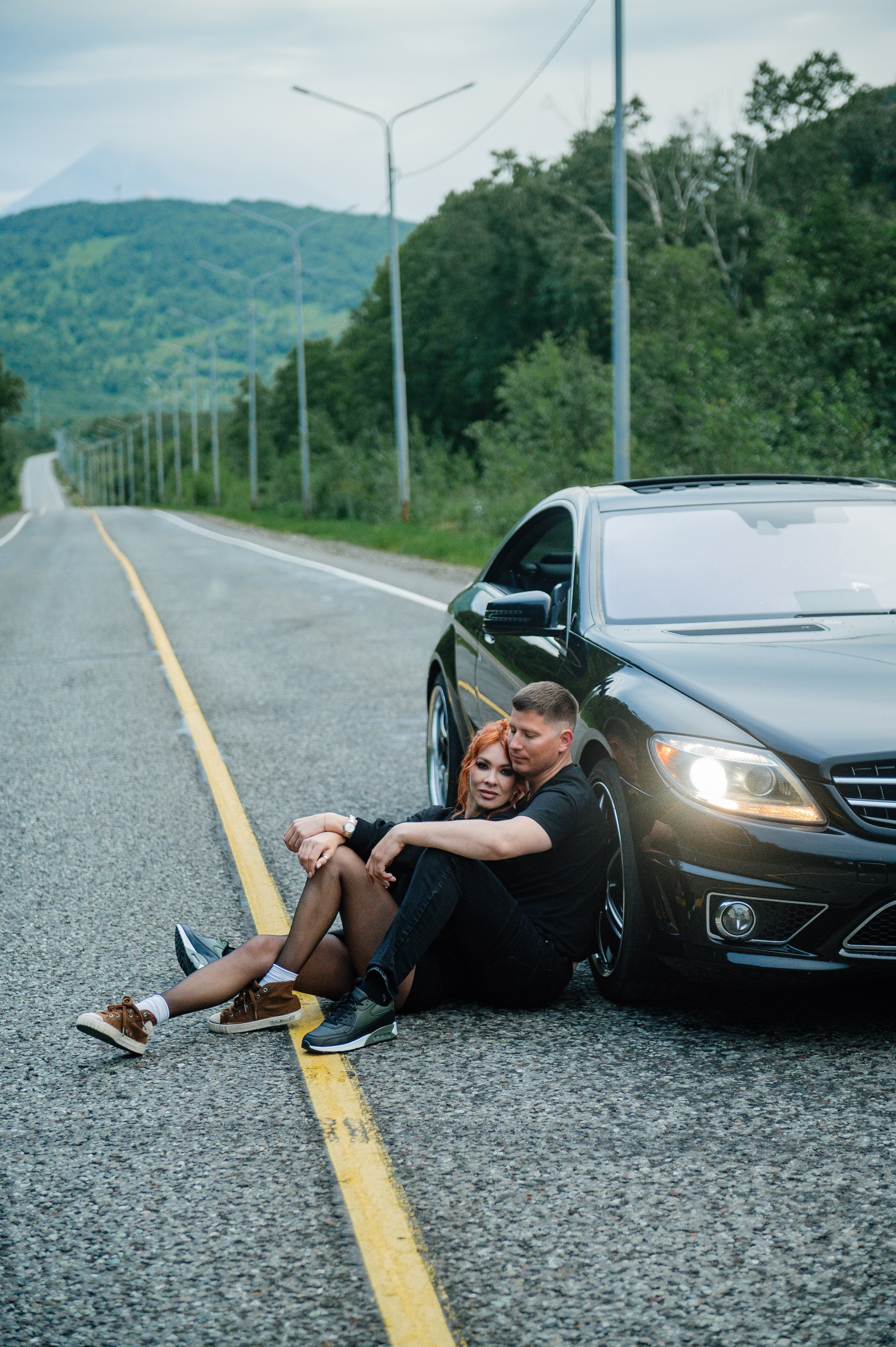 Пара сидит на асфальте у чёрного автомобиля на пустой дороге. Вокруг — зелёные холмы, атмосфера спокойная, романтичная.