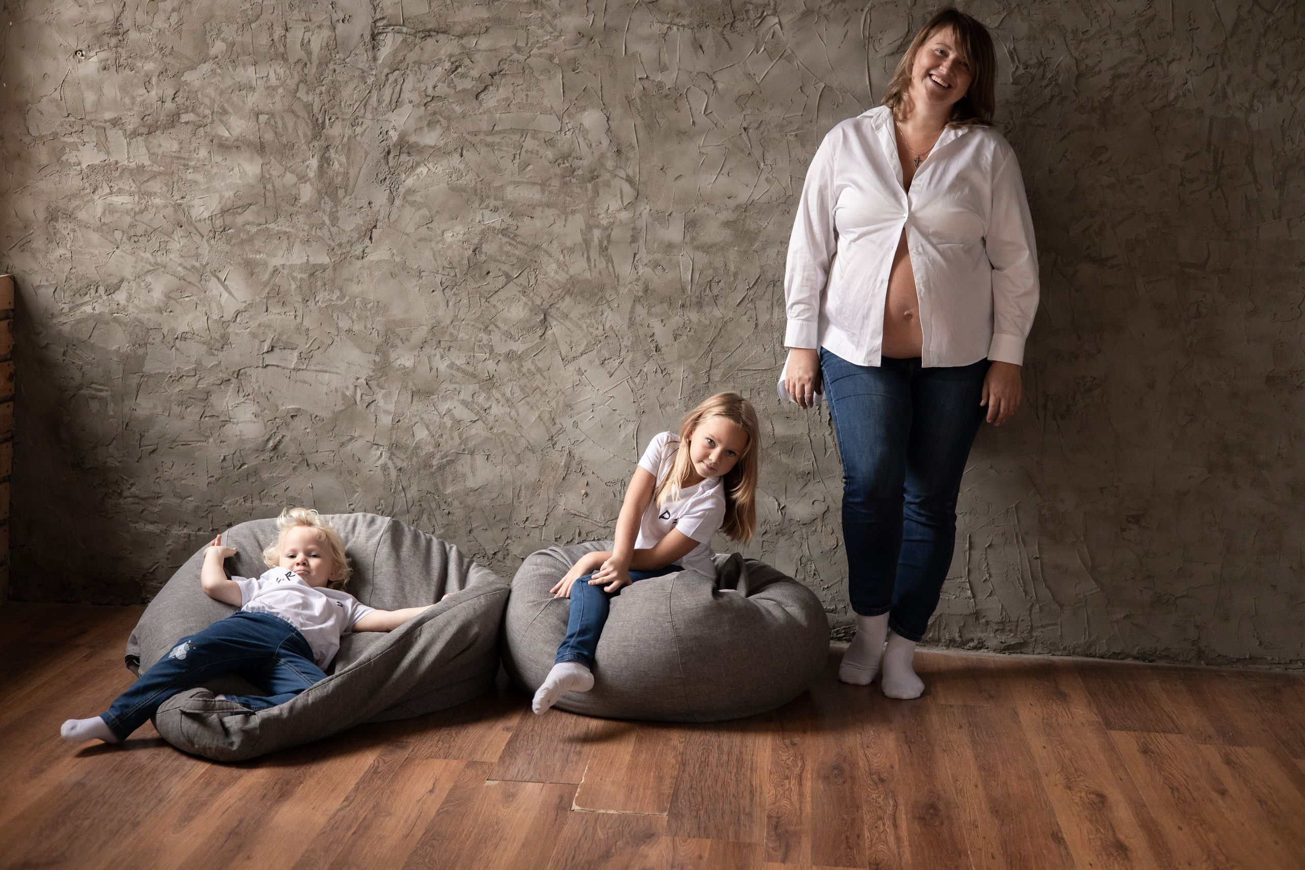 Беременная мама и двое её детей. Дети лежат на мягких креслах, мама стоит у стены. Семейная фотосессия Астрахань.