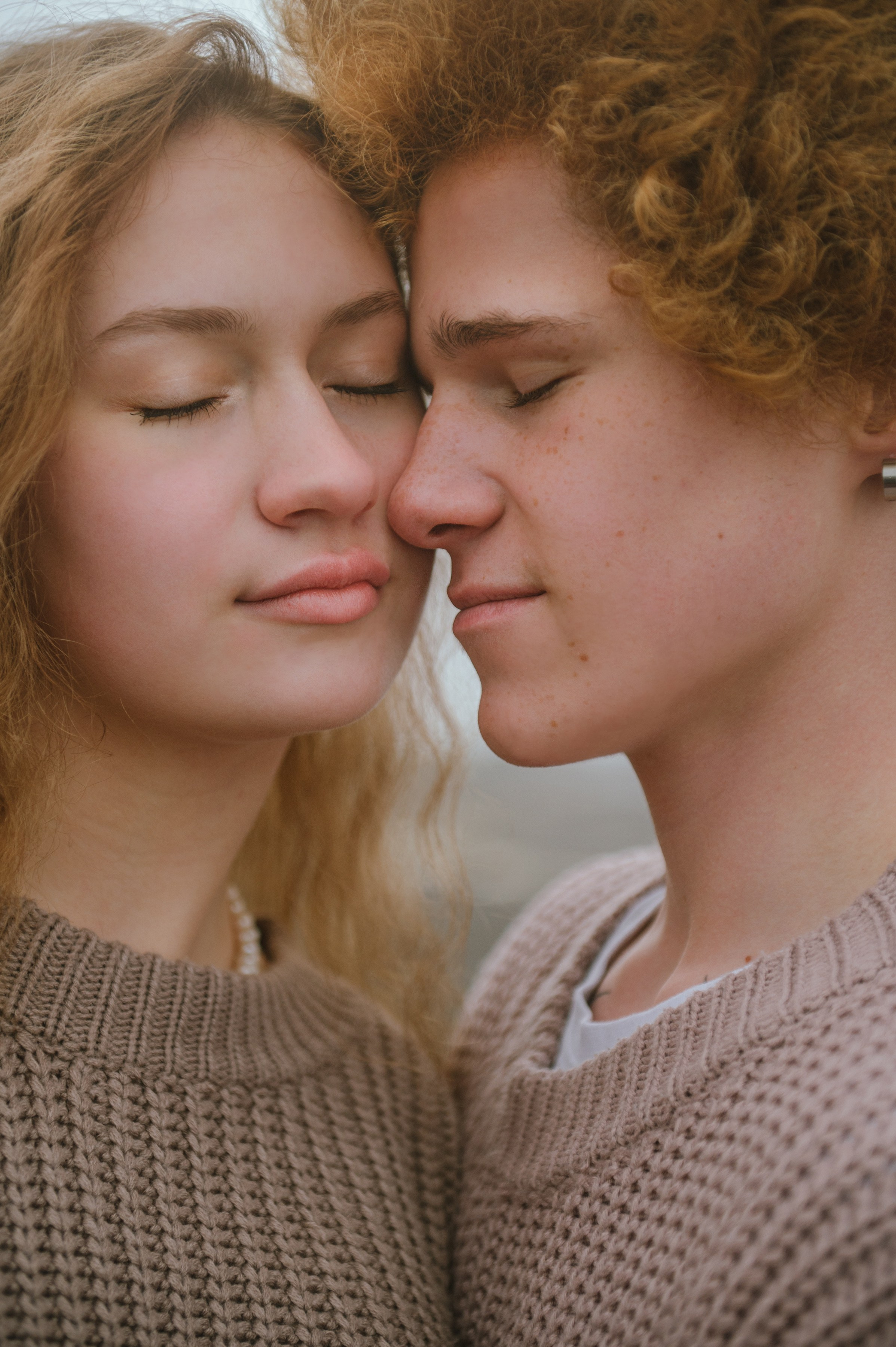 Парень с кудрявыми волосами и девушка с длинными светлыми волосами стоят близко, лбы соприкасаются, глаза закрыты. Атмосфера нежная, романтичная.