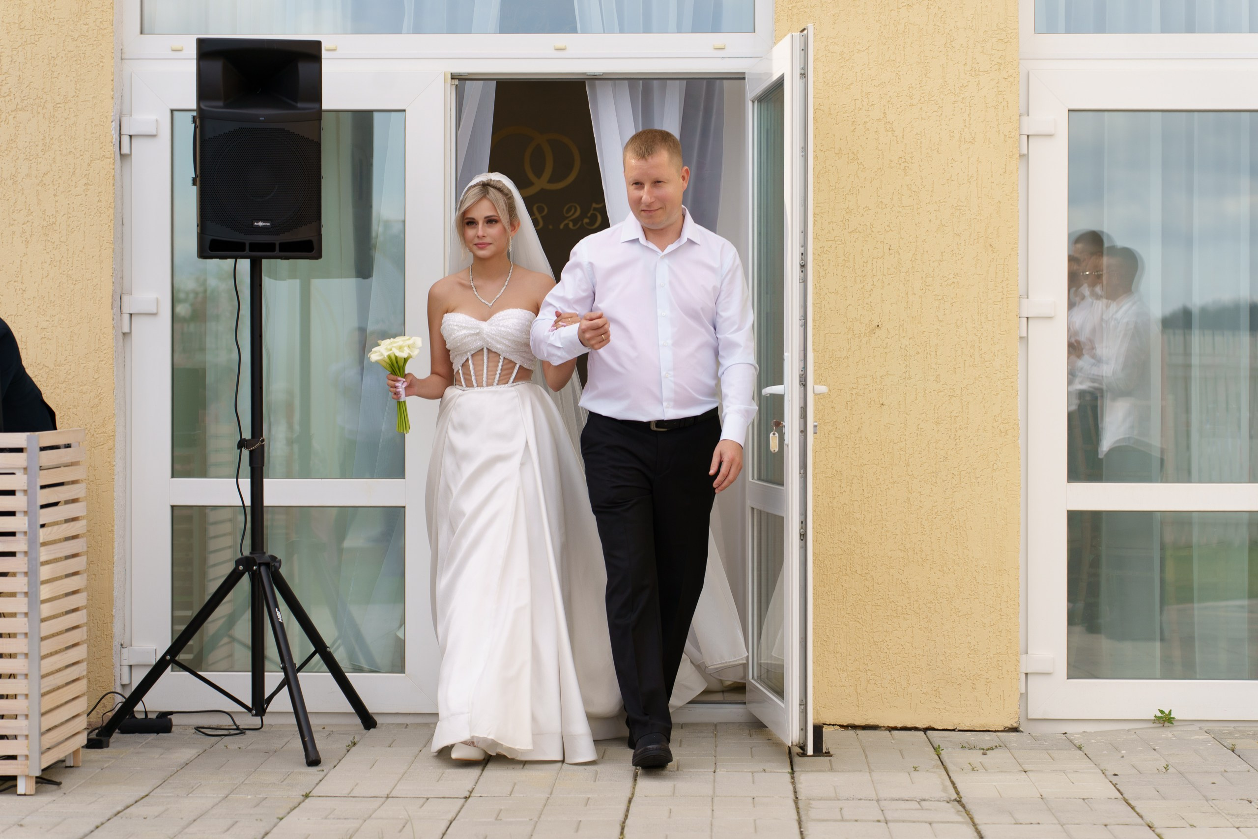 Трогательный момент выхода невесты в изысканном белом платье с букетом нежных калл и отца невесты в элегантной белой рубашке и чёрных брюках из дверей зала, украшенного свадебной символикой | фотограф для выездной регистрации свадьбы Екатеринбург