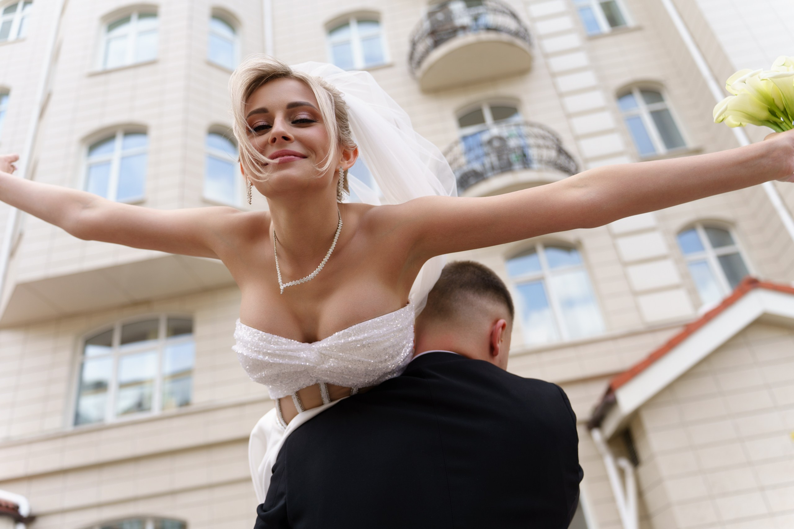 Невеста в роскошном свадебном платье с фатой сидит на плечах жениха, её лицо озарено улыбкой, а руки раскинуты в жесте безграничного счастья | фотограф для традиционных свадеб Екатеринбург