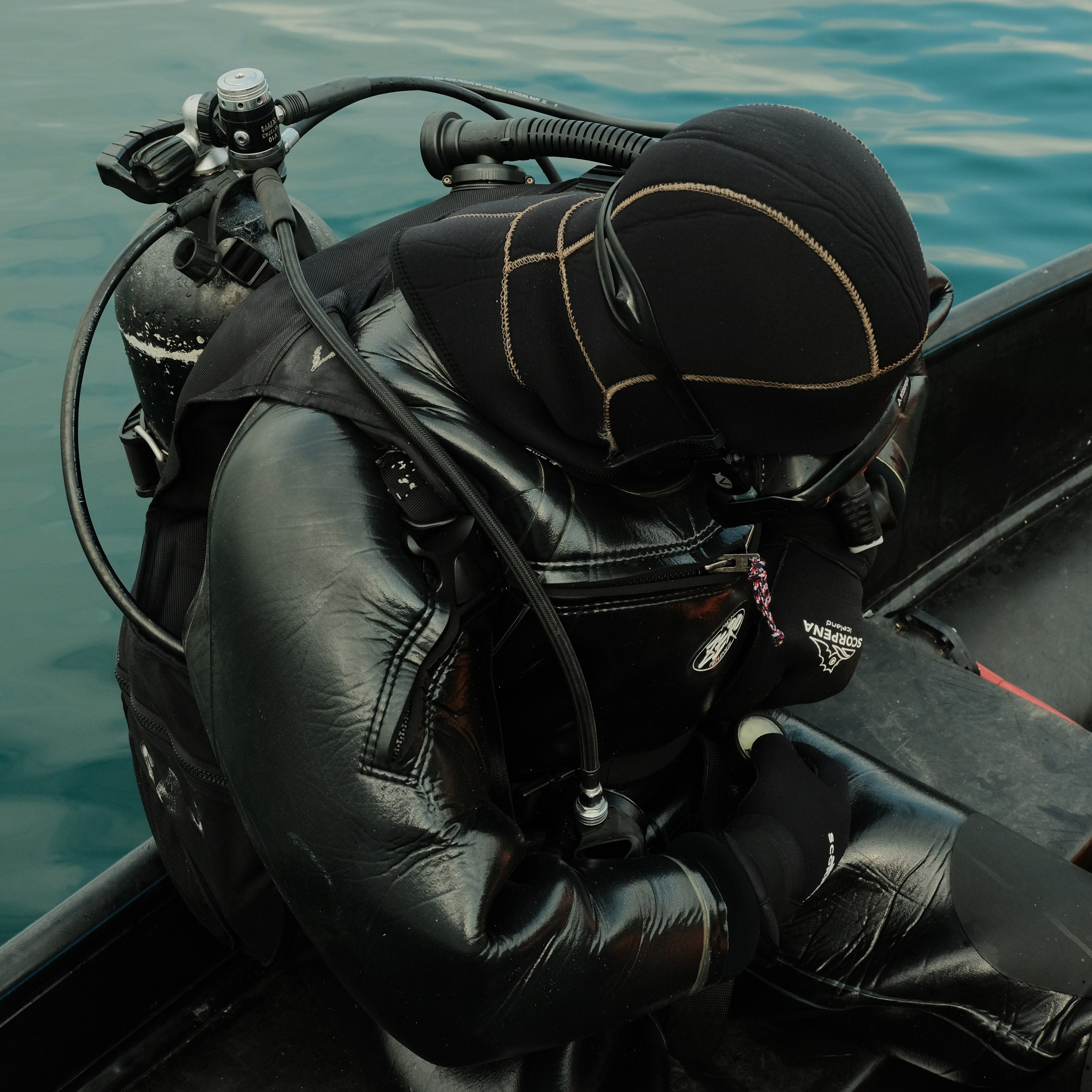 A scuba diver in a black wetsuit and hood sits on the edge of a boat, adjusting equipment near calm blue water.