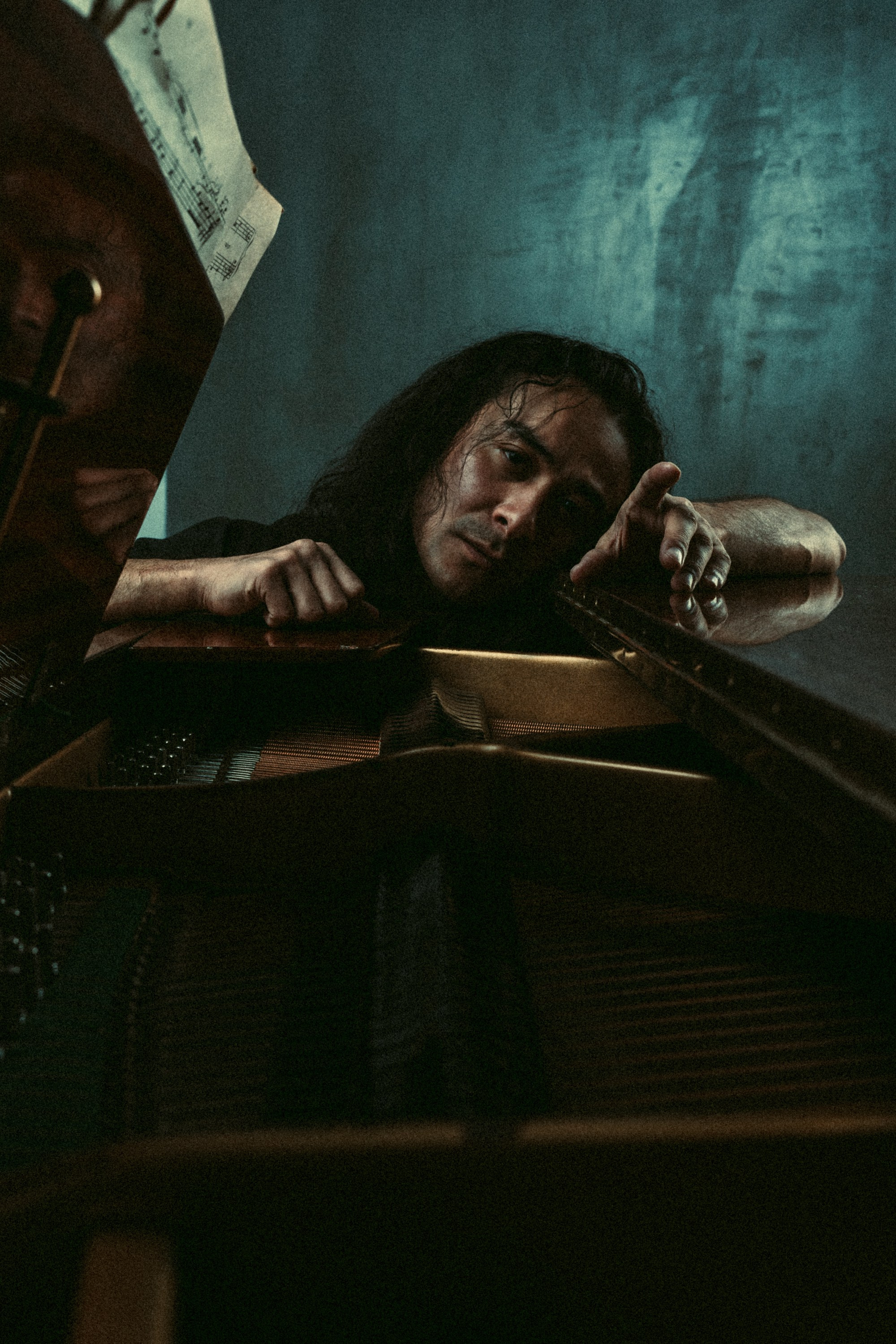 Мужчина с длинными тёмными волосами лежит на рояле, его лицо отражает усталость и эмоции в затемнённой комнате.