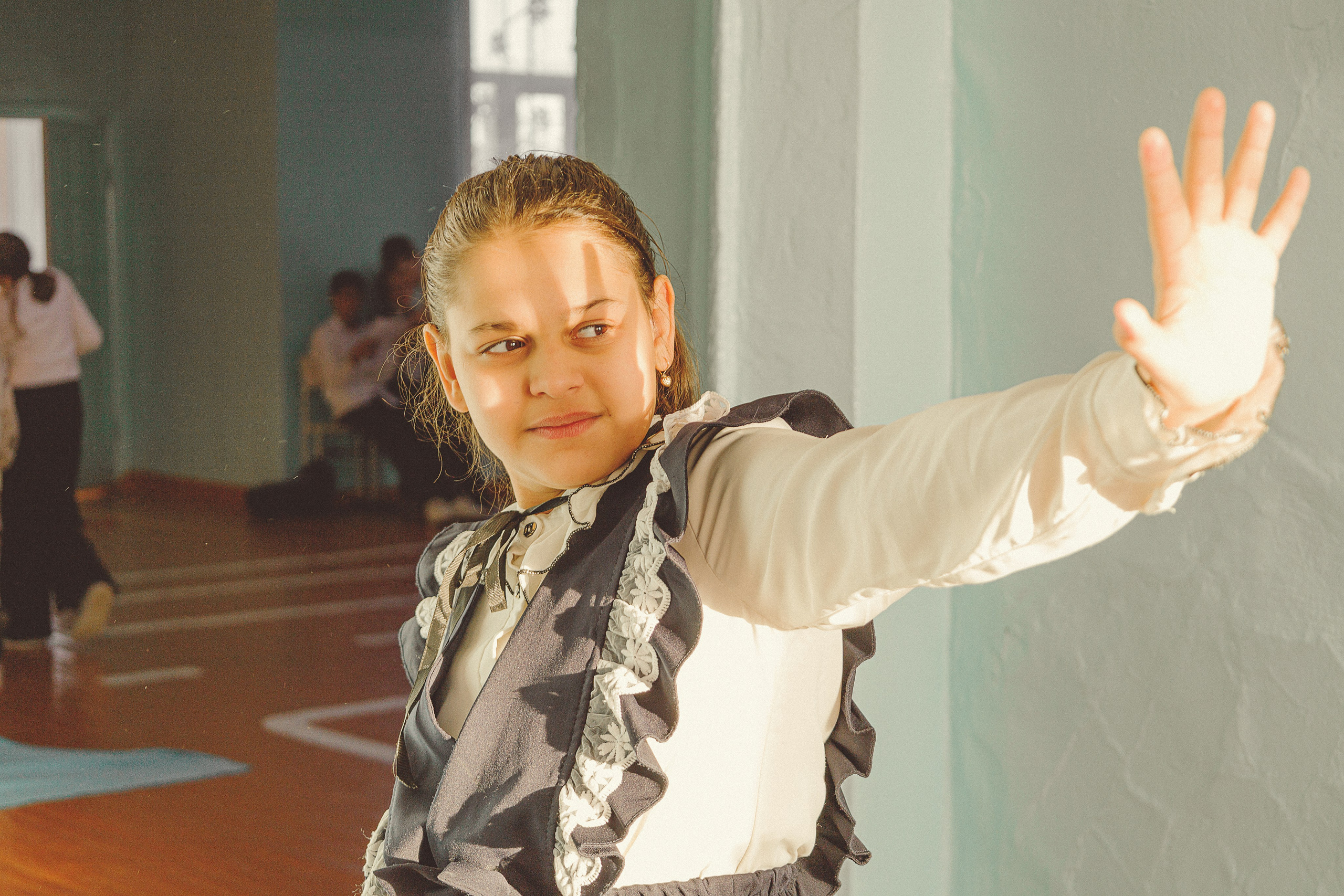 Девочка в школьной форме уверенно позирует с вытянутой вперед рукой, солнечный свет отбрасывает тени на ее лицо, что говорит о решимости.