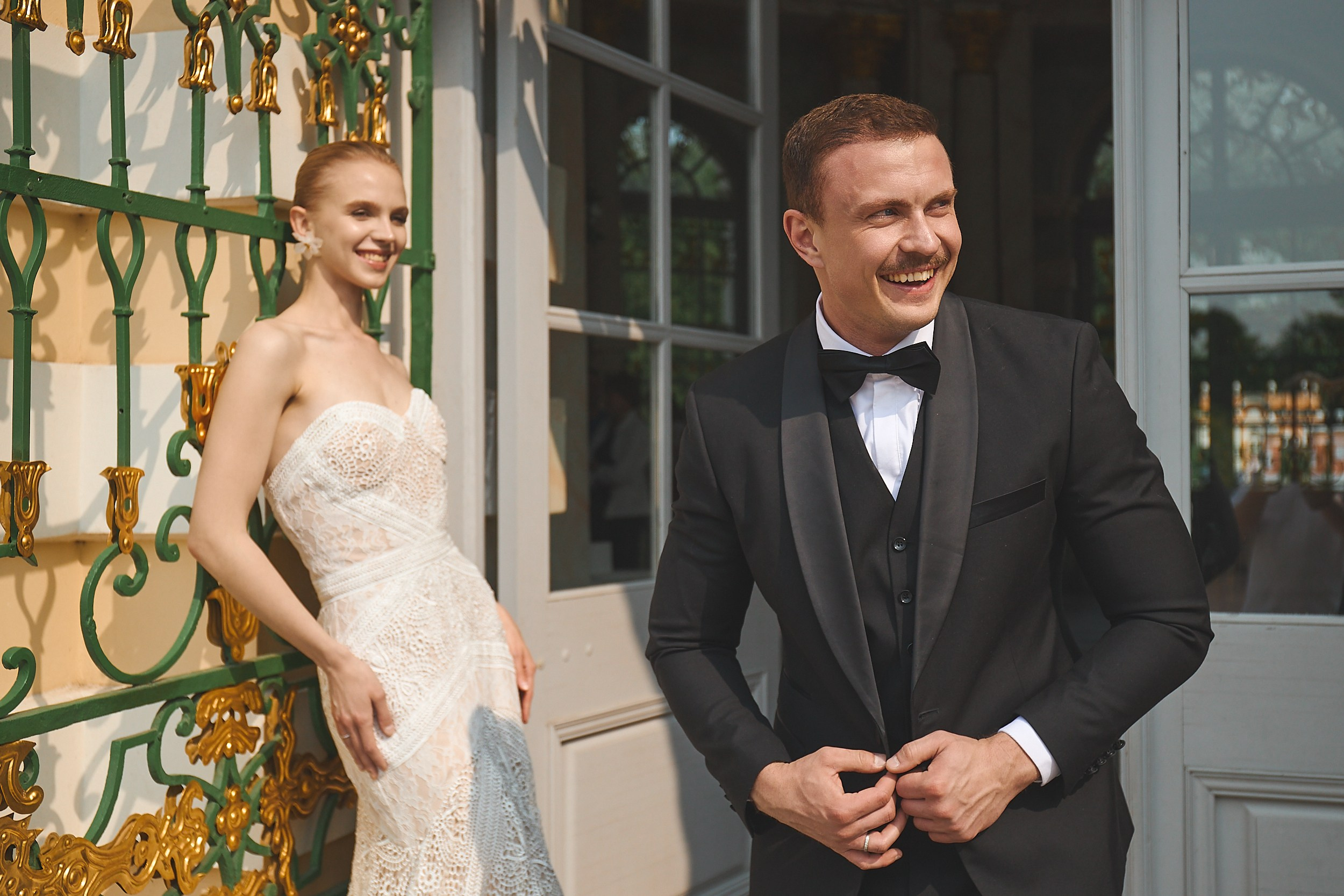 Жених в черном смокинге улыбается рядом со своей невестой в белом платье на фоне красивого дома в парке Кусково