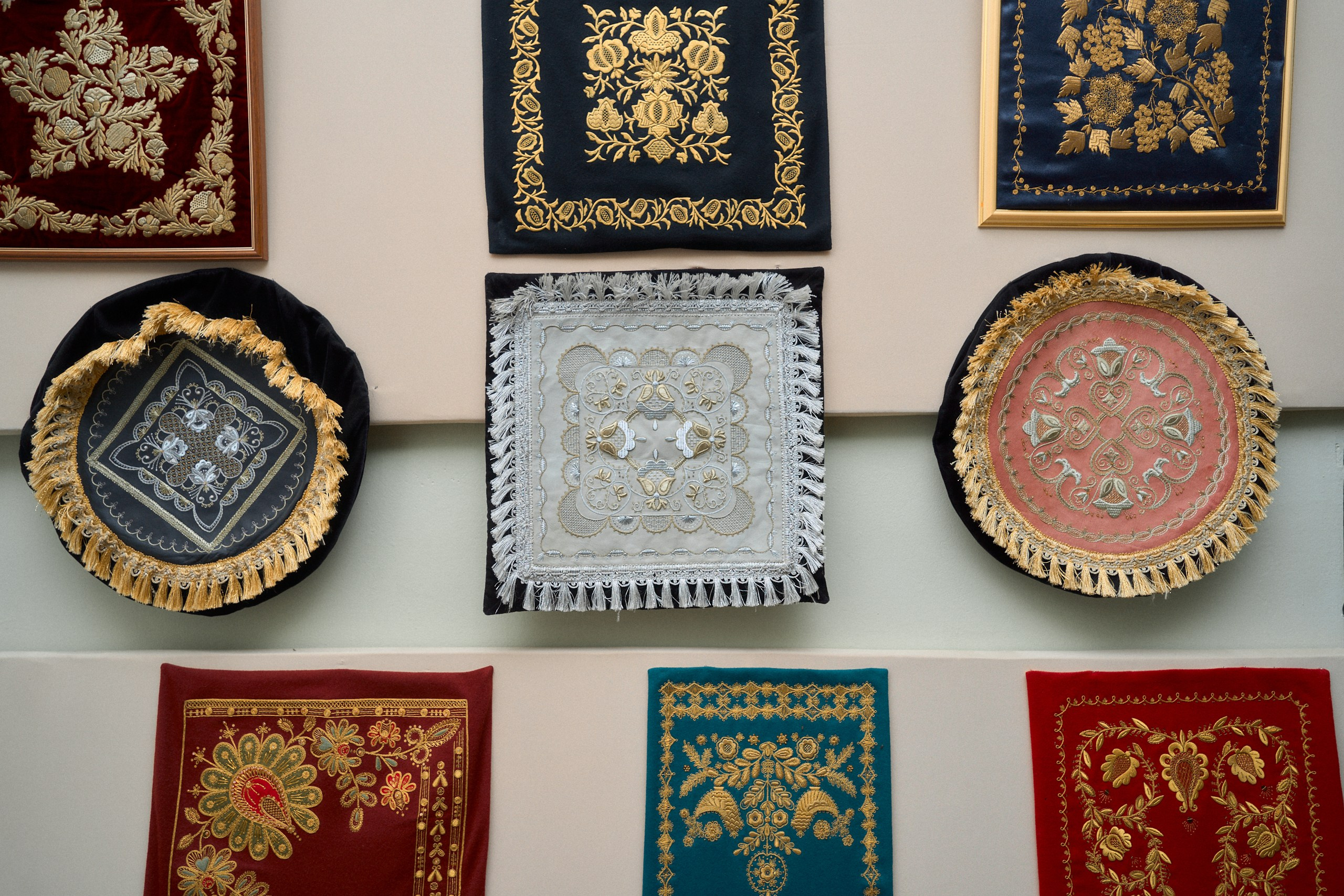 Экспозиция в Музее торжокских золотошвей в Торжке, представляющая разнообразные образцы золотого шитья, включая вышитые золотом подушки и ткани с традиционными узорами, выставленные на стене
