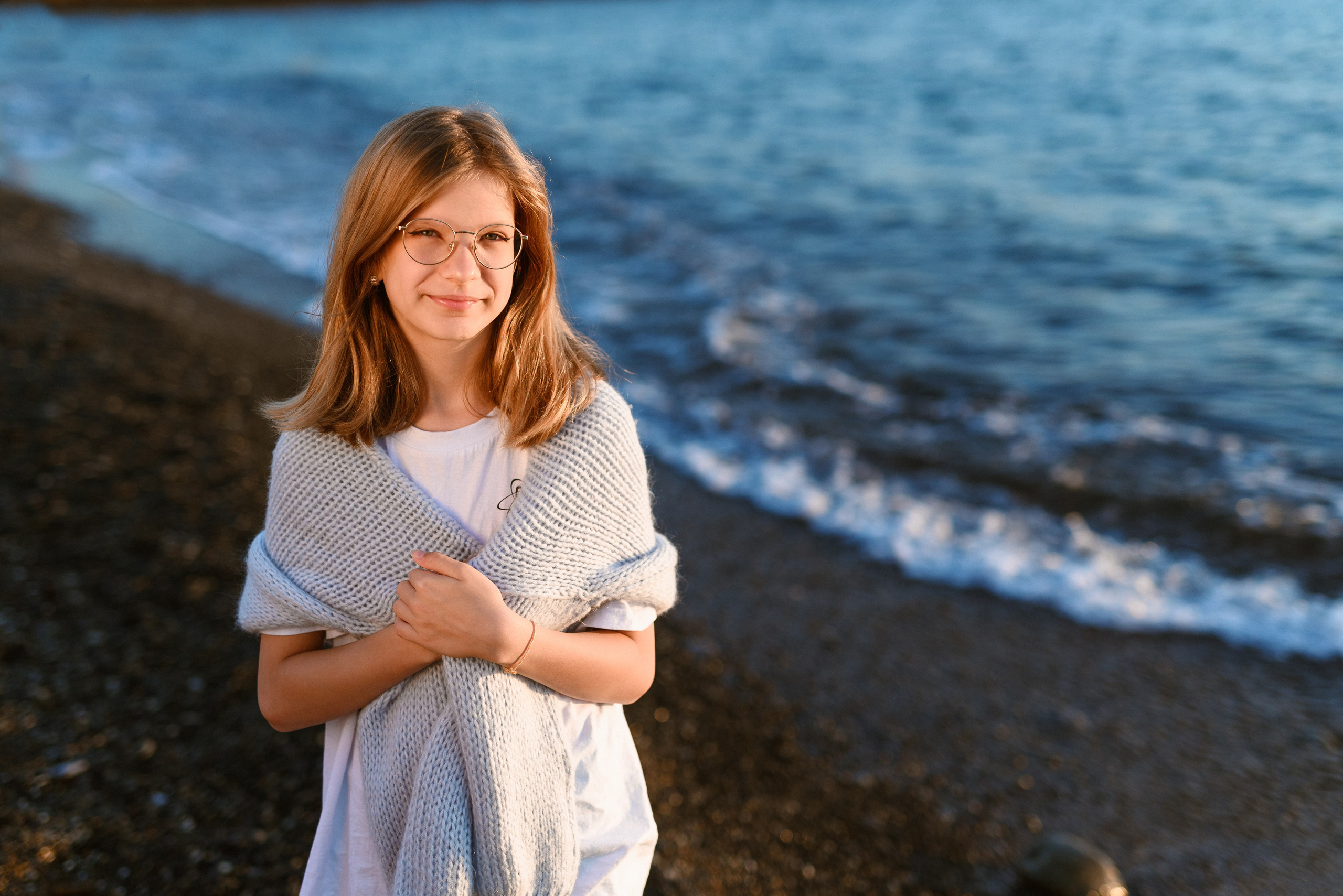 Девочка в очках стоит смотря в камеру на фоне линии прибоя
