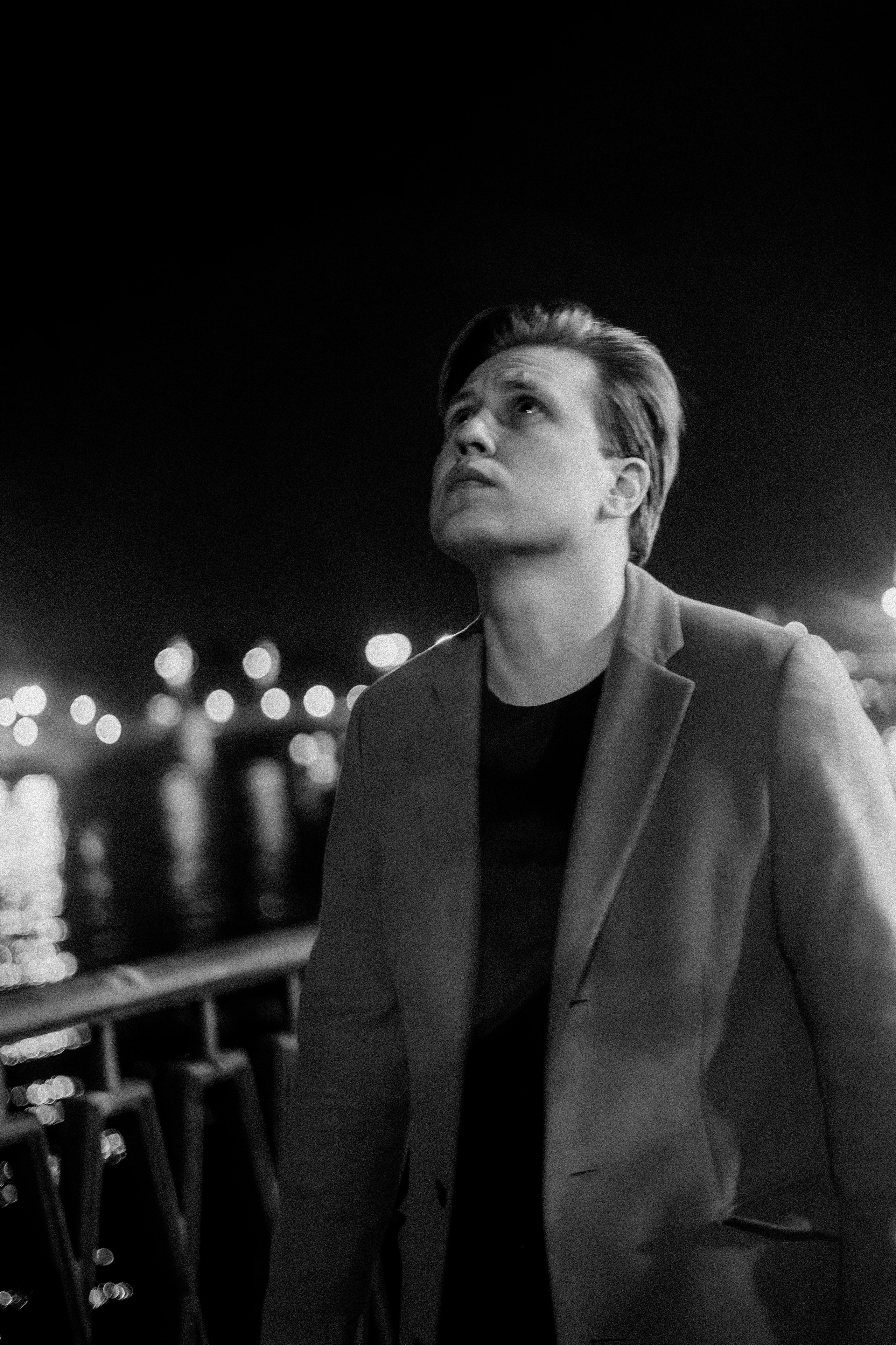 Мужчина в пальто смотрит вверх, идёт по набережной ночью.