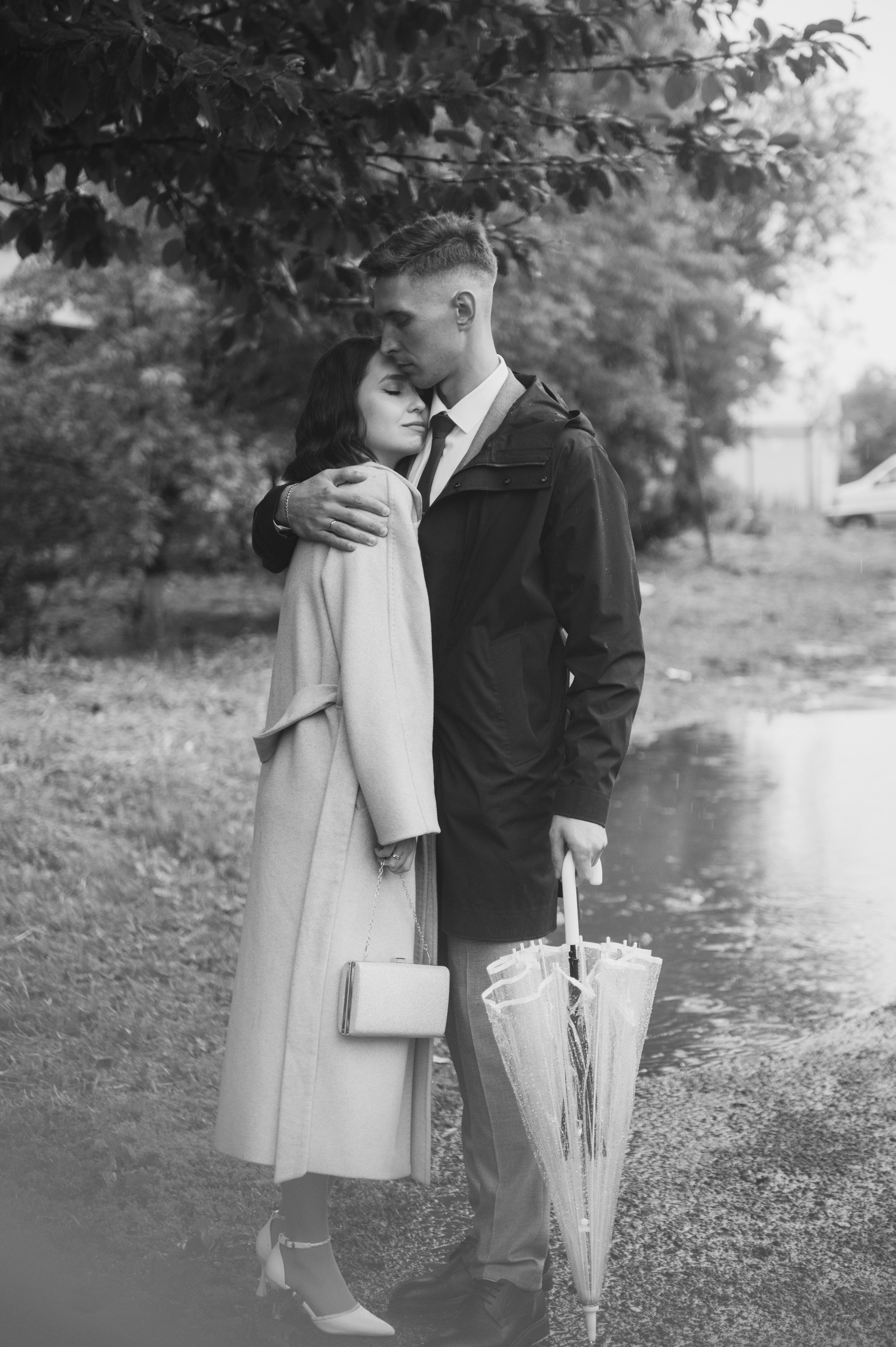 Парень в плаще с прозрачным зонтом обнимает девушку в светлом пальто у пруда. Атмосфера нежная, романтичная, немного дождливая.