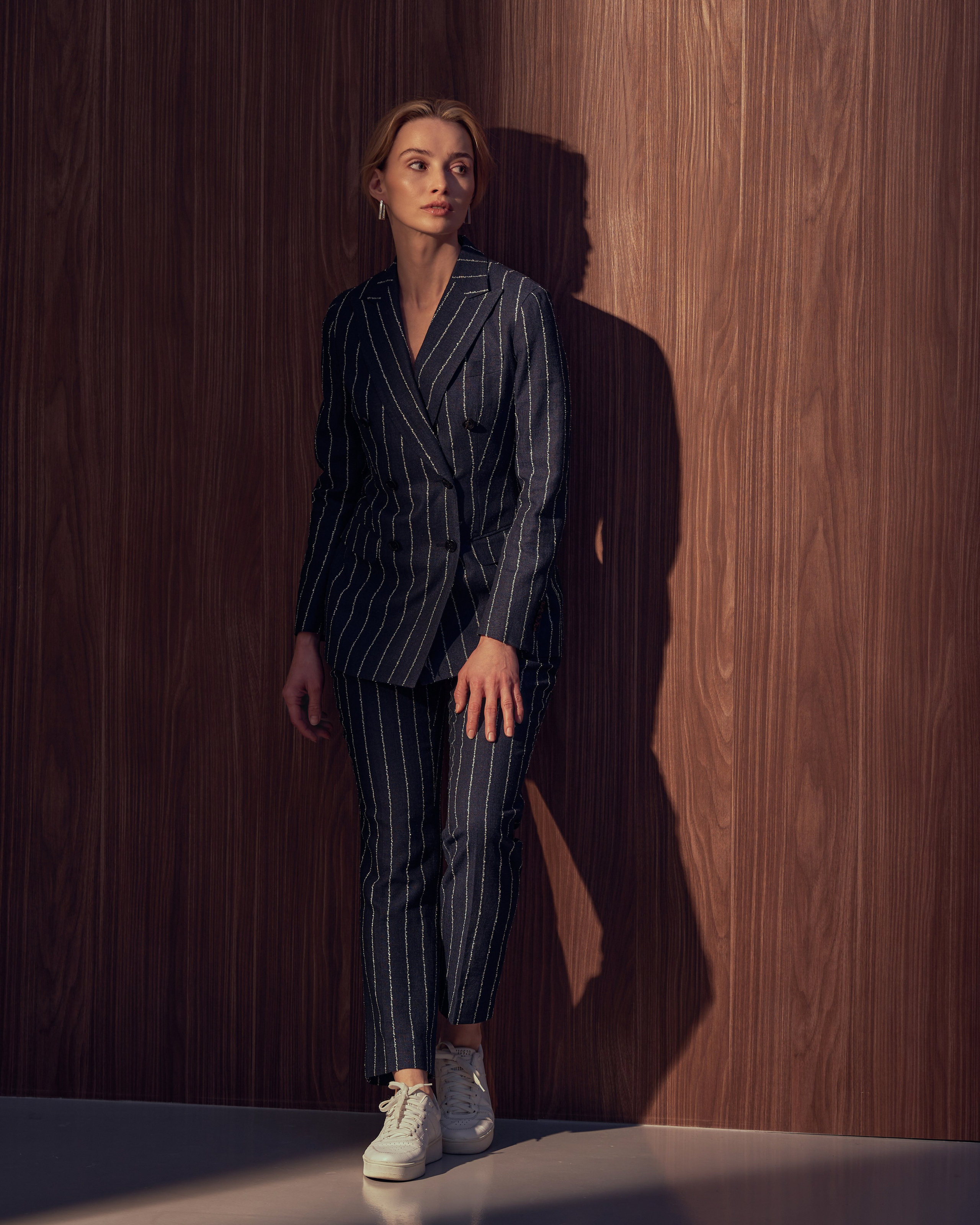 Съемка одежды, девушка на фоне деревянной стены в брючном костюме в полоску и кедах, девушка в костюме и кедах.