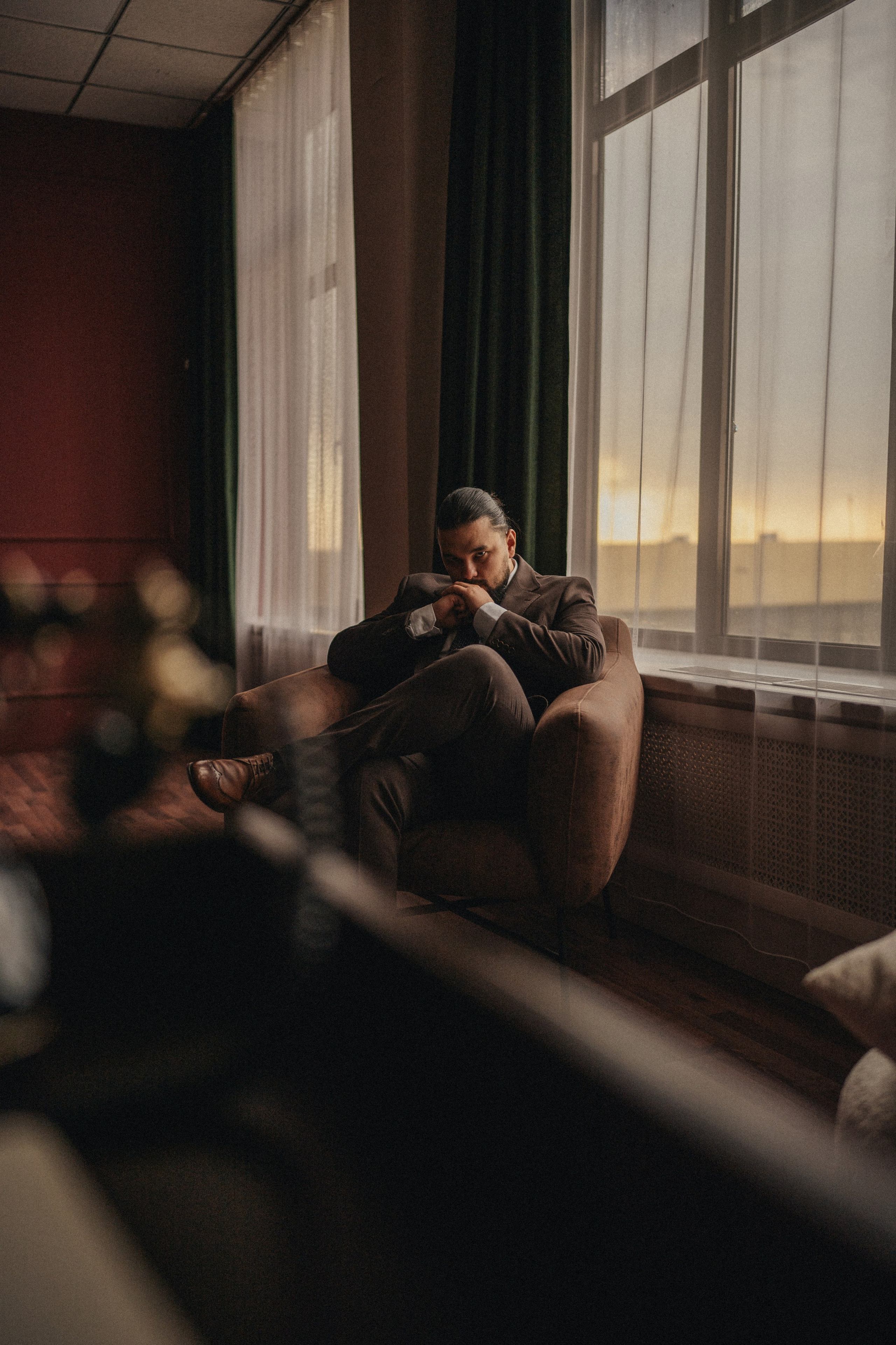 Мужчина в коричневом костюме задумчиво сидит в кресле перед окном на закат