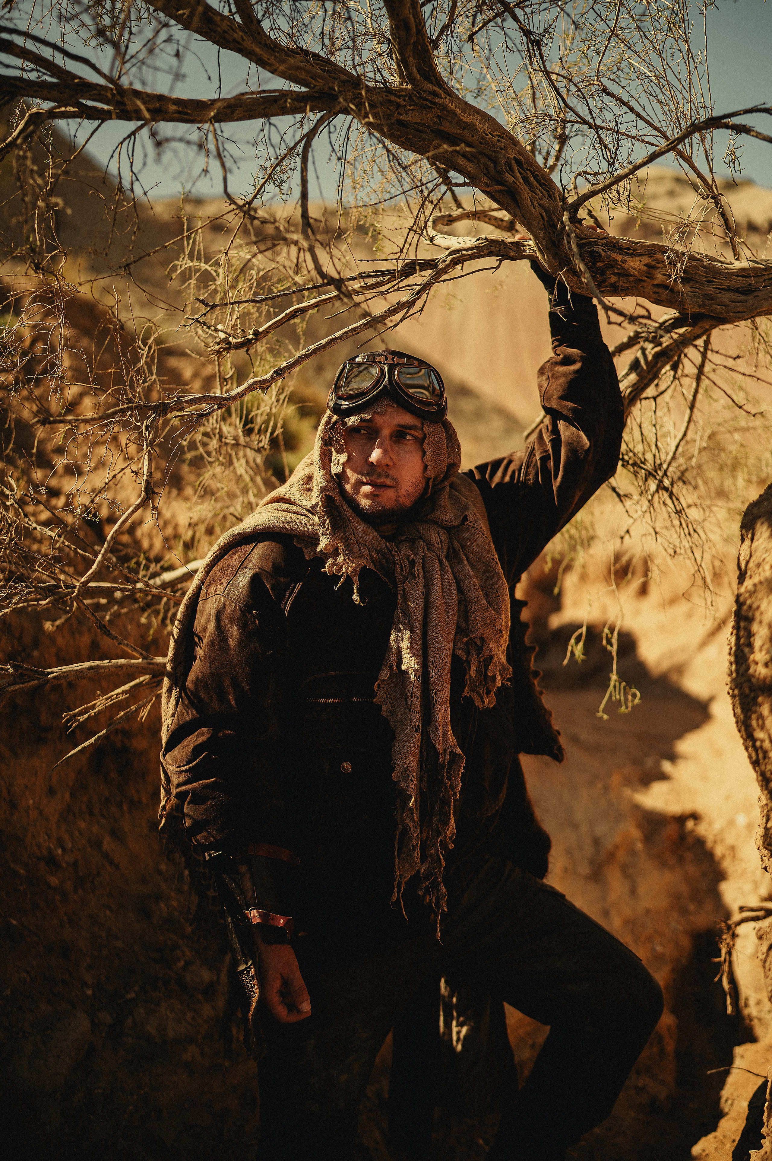 Мужчина в защитных очках и шарфе стоит под засохшим деревом в пустыне с жёлтым песком, фотосессия в стиле Дюна