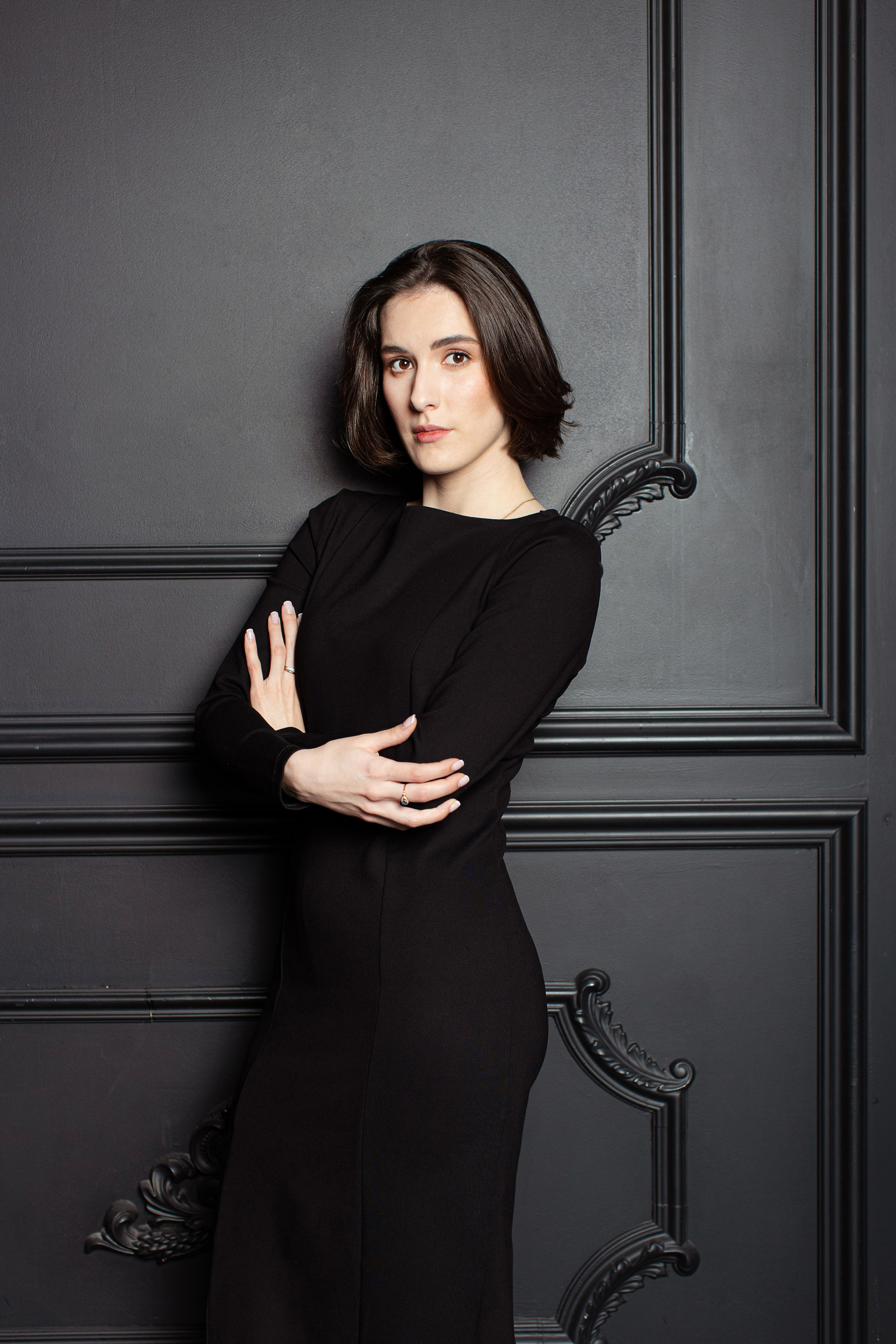 Девушка брюнетка в черном платье стоит у темной стены