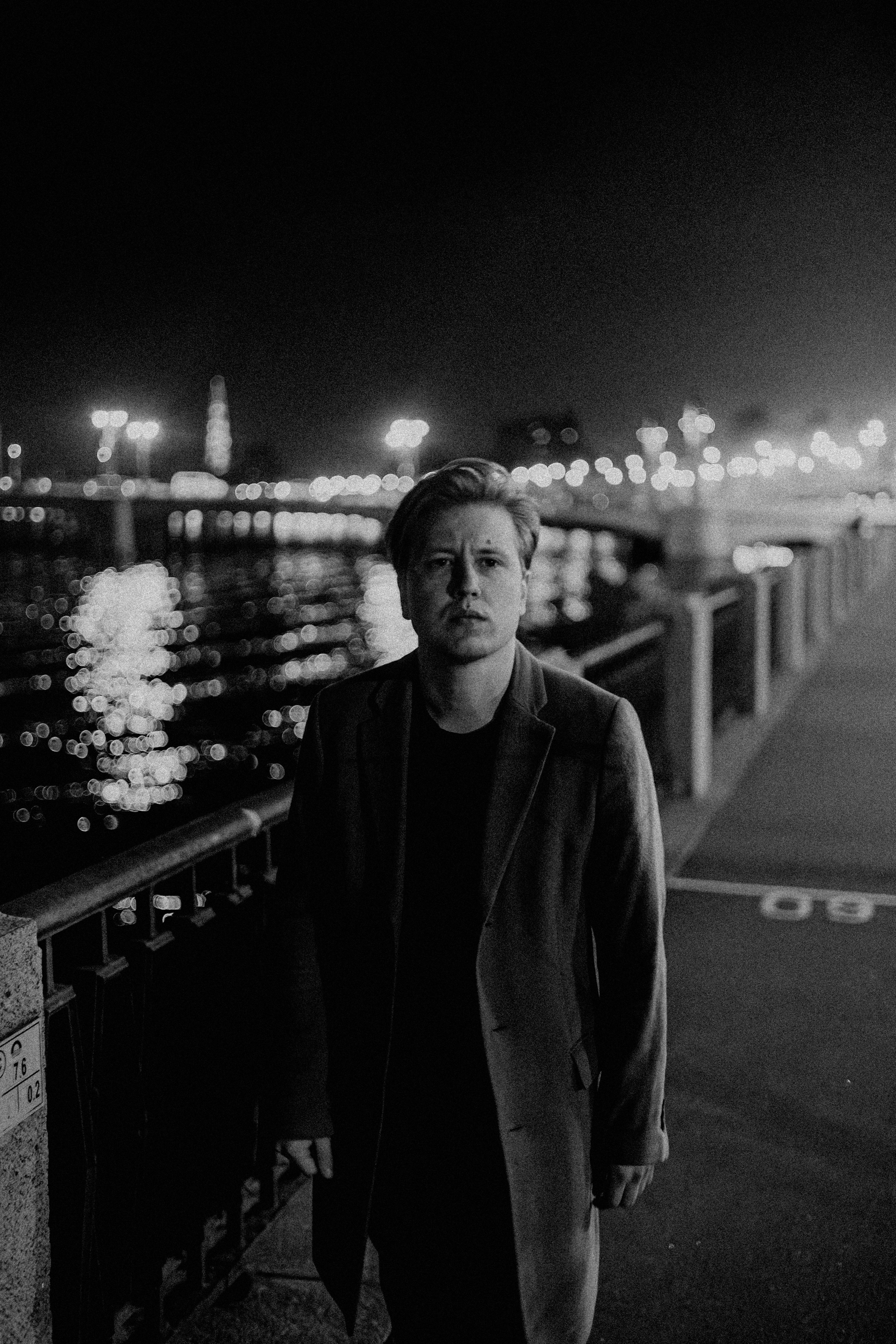 Молодой человек в черной футболке и пальто идёт по набережной один.