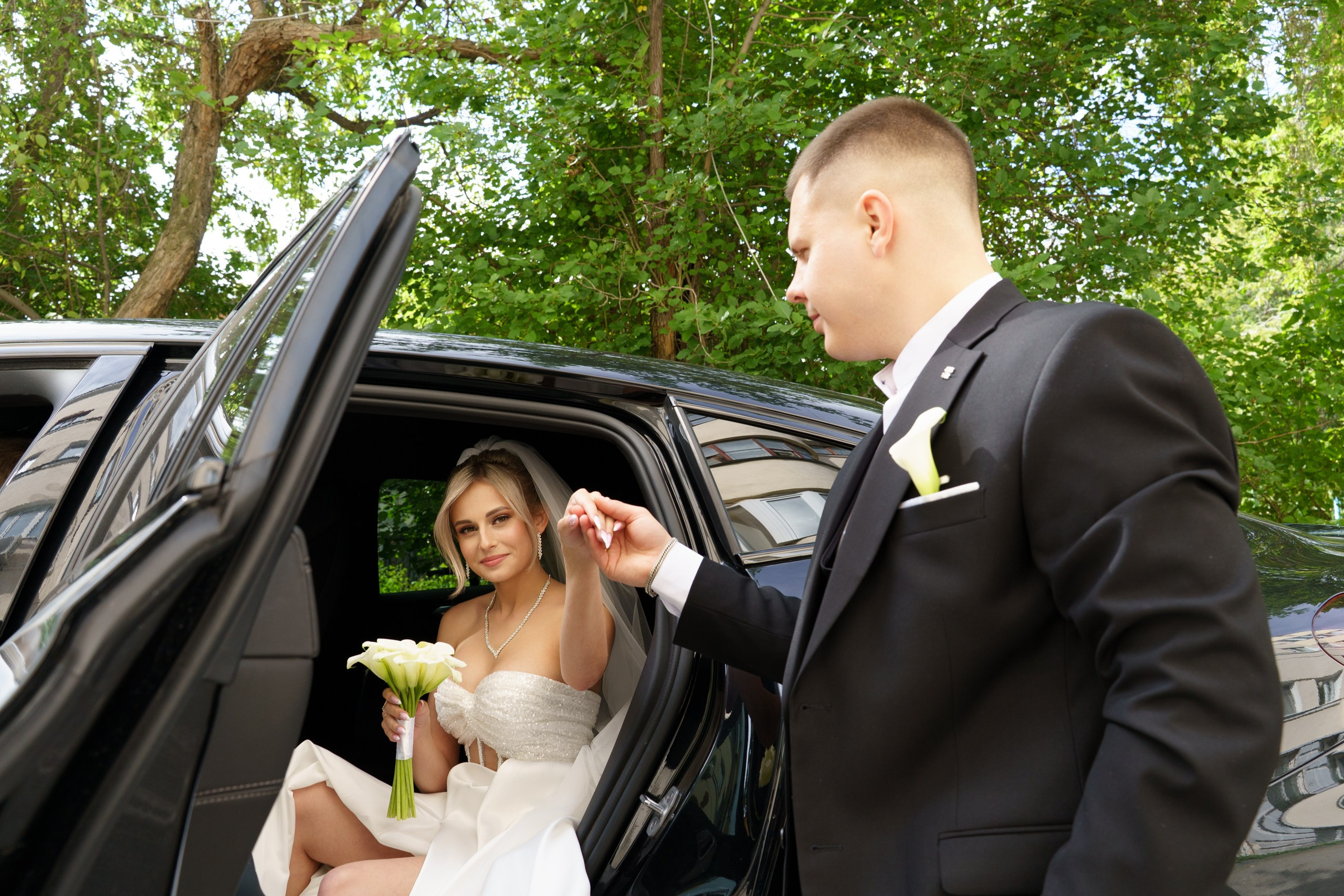 Сцена из свадебного дня: жених в строгом костюме с бутоньеркой и невеста в белом платье с букетом калл. Жених помогает невесте выйти из автомобиля на фоне зелёных деревьев | фотограф для инстаграм-свадьбы Екатеринбург