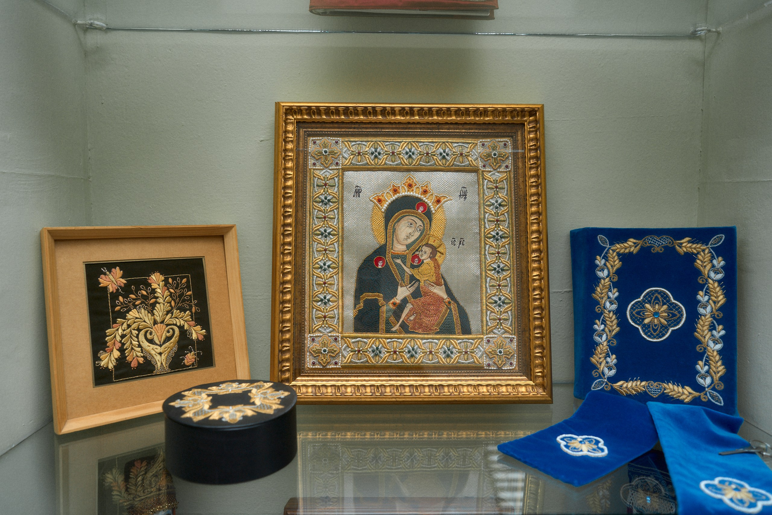 Витрина с образцами торжокского золотного шитья, включающая икону в золотой раме, вышитую золотыми нитями, а также другие декоративные изделия с традиционными узорами на синем и чёрном бархате.