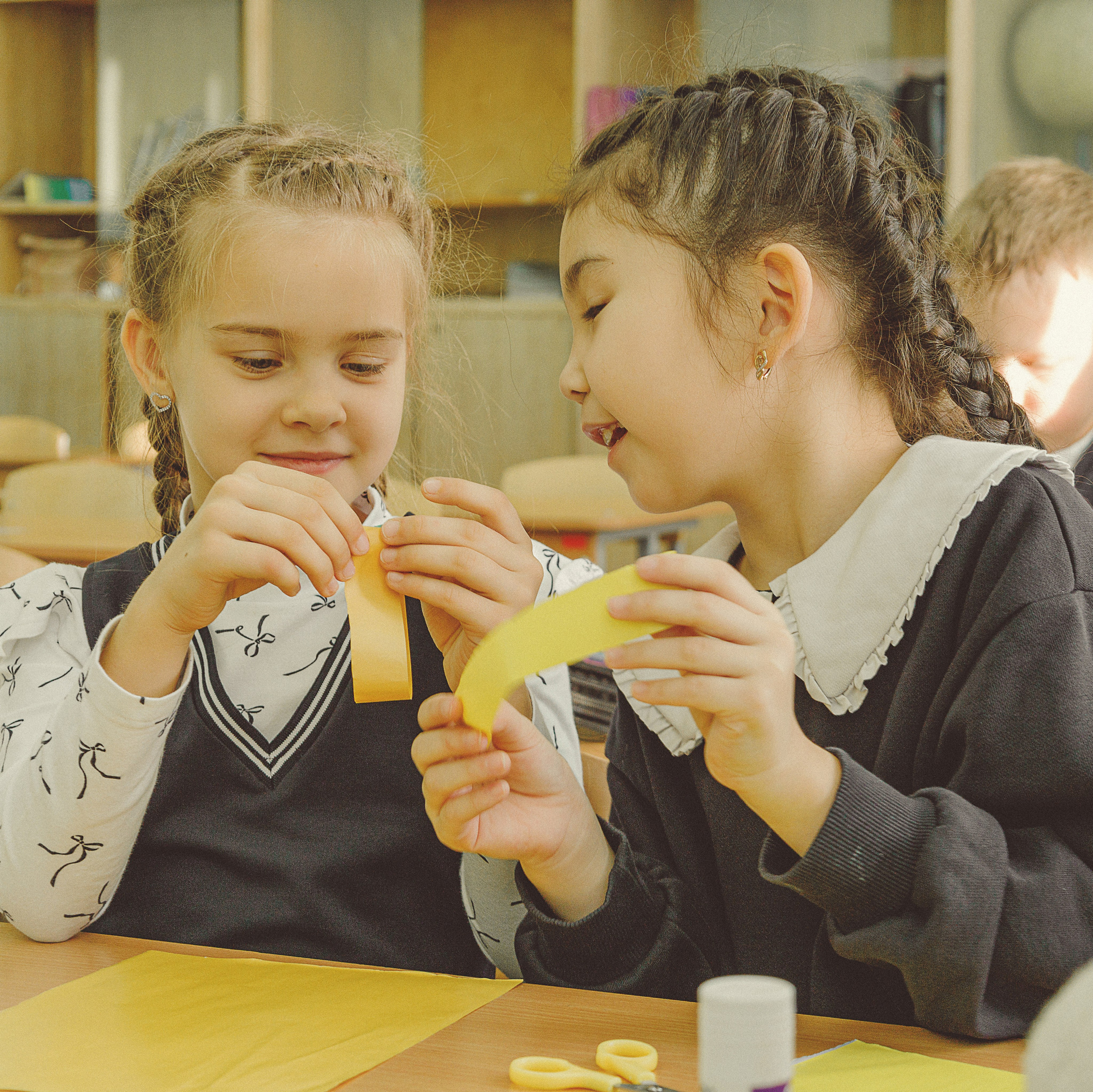 Две девочкии в школьной форме, с заплетенными в косички волосами, радостно работают вместе над поделкой в ярко освещенном классе. На заднем плане видны другие дети.