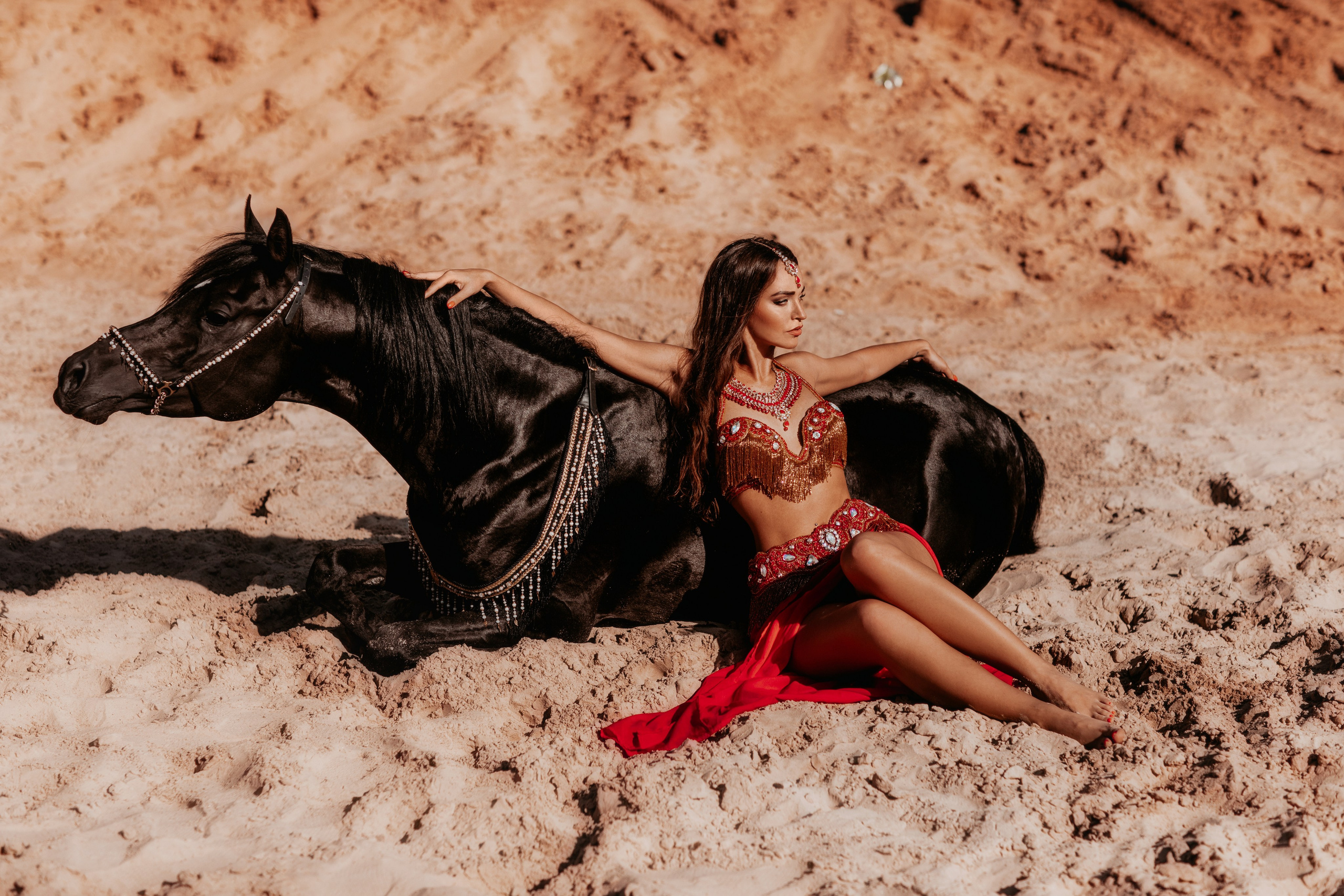 "Девушка в ярком восточном платье нежно сидит рядом с лежащей лошадью на мягкой траве. Спокойное выражение лица и расслабленная поза животного создают атмосферу умиротворения. Эта фотография идеально подходит для клиентов в Москве и области, желающих запечатлеть уникальные моменты с животными."
