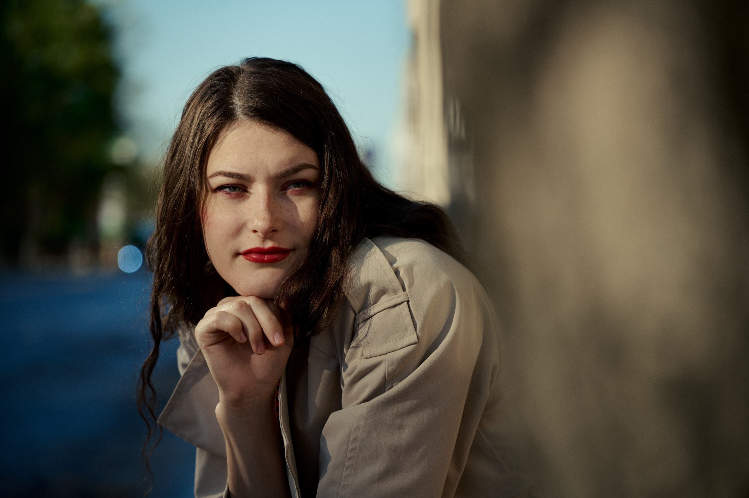 Эмоциональный уличный портрет: женщина в светлом пальто и красной помаде смотрит в камеру, опираясь на стену.