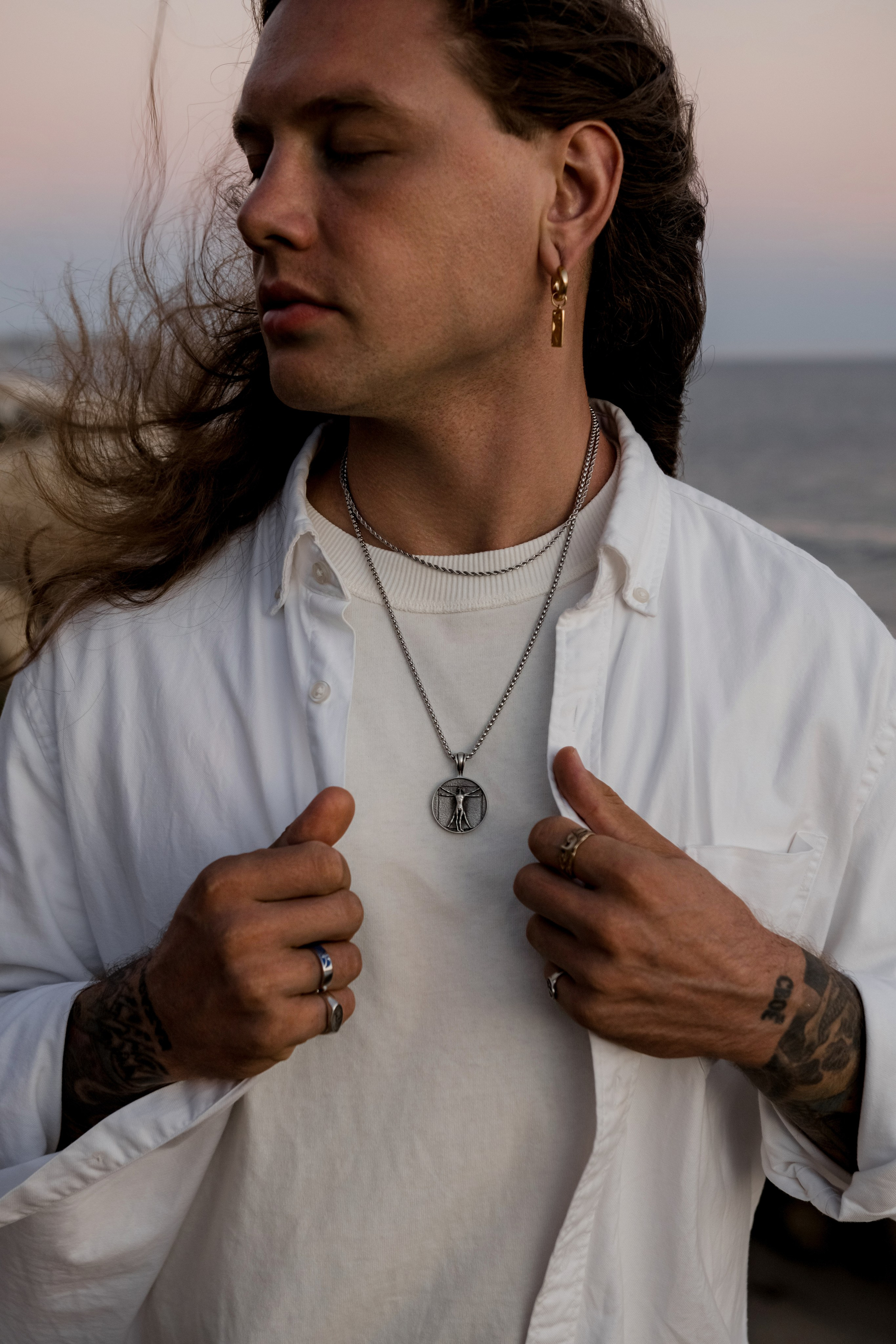 Male model adjusting his silver necklace on a beach, blending rugged and modern elegance.