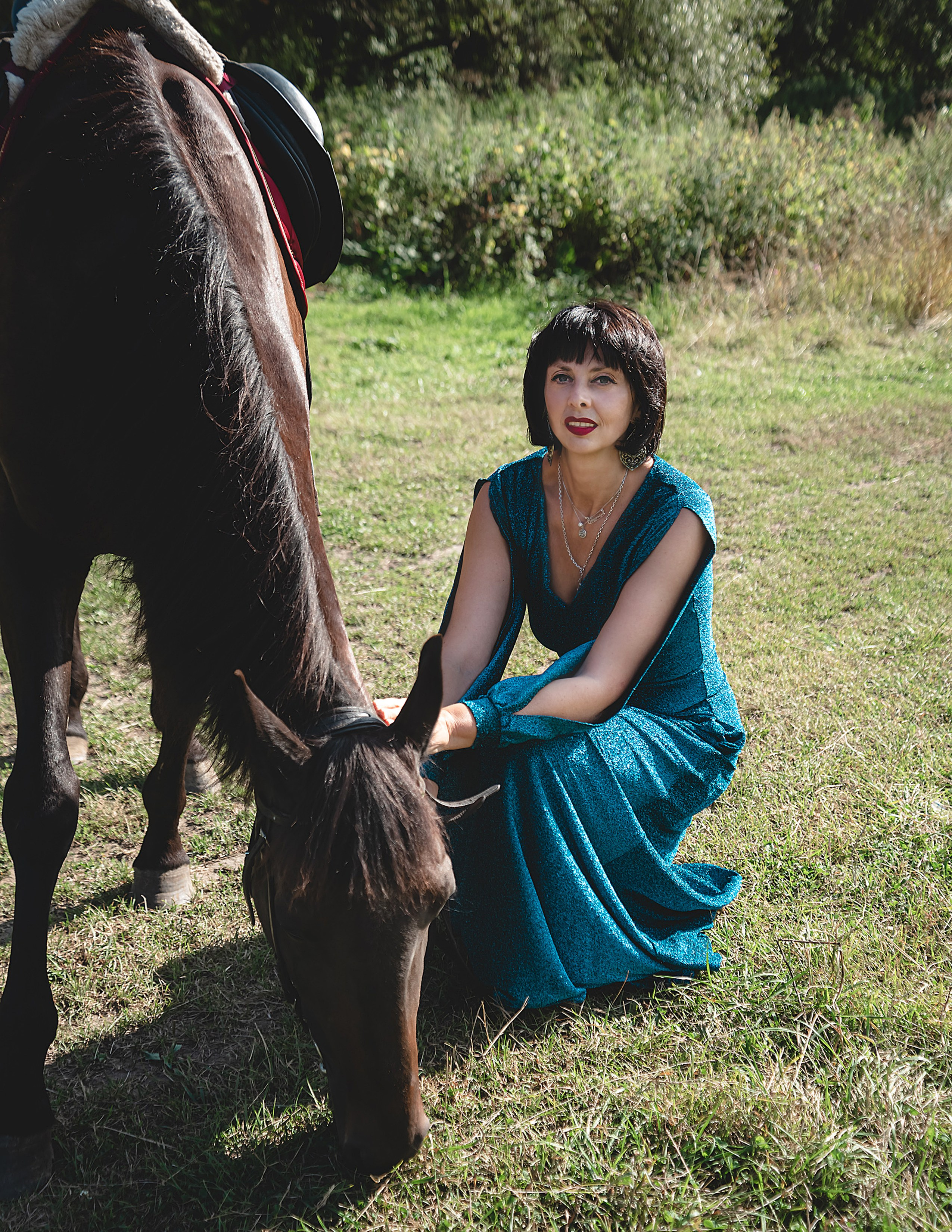 На фото женщина в зеленом платье сидит около  лошади и кормит ее.