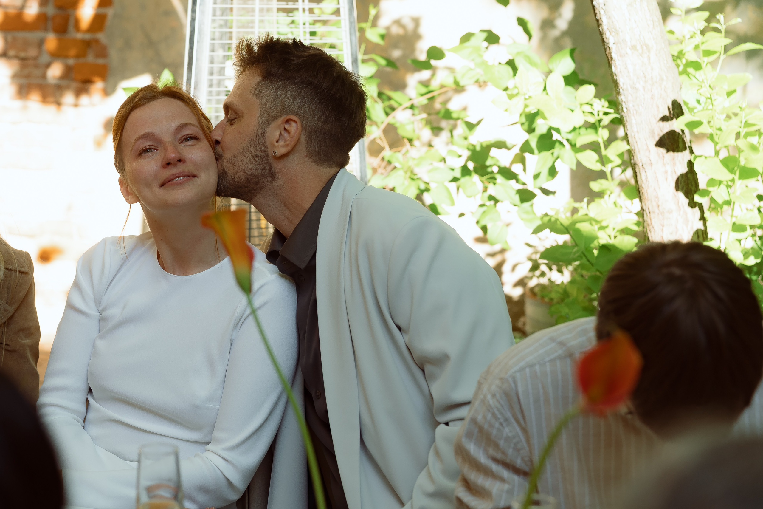 Евгений целует Марию в щёку у столика под зелёными ветвями