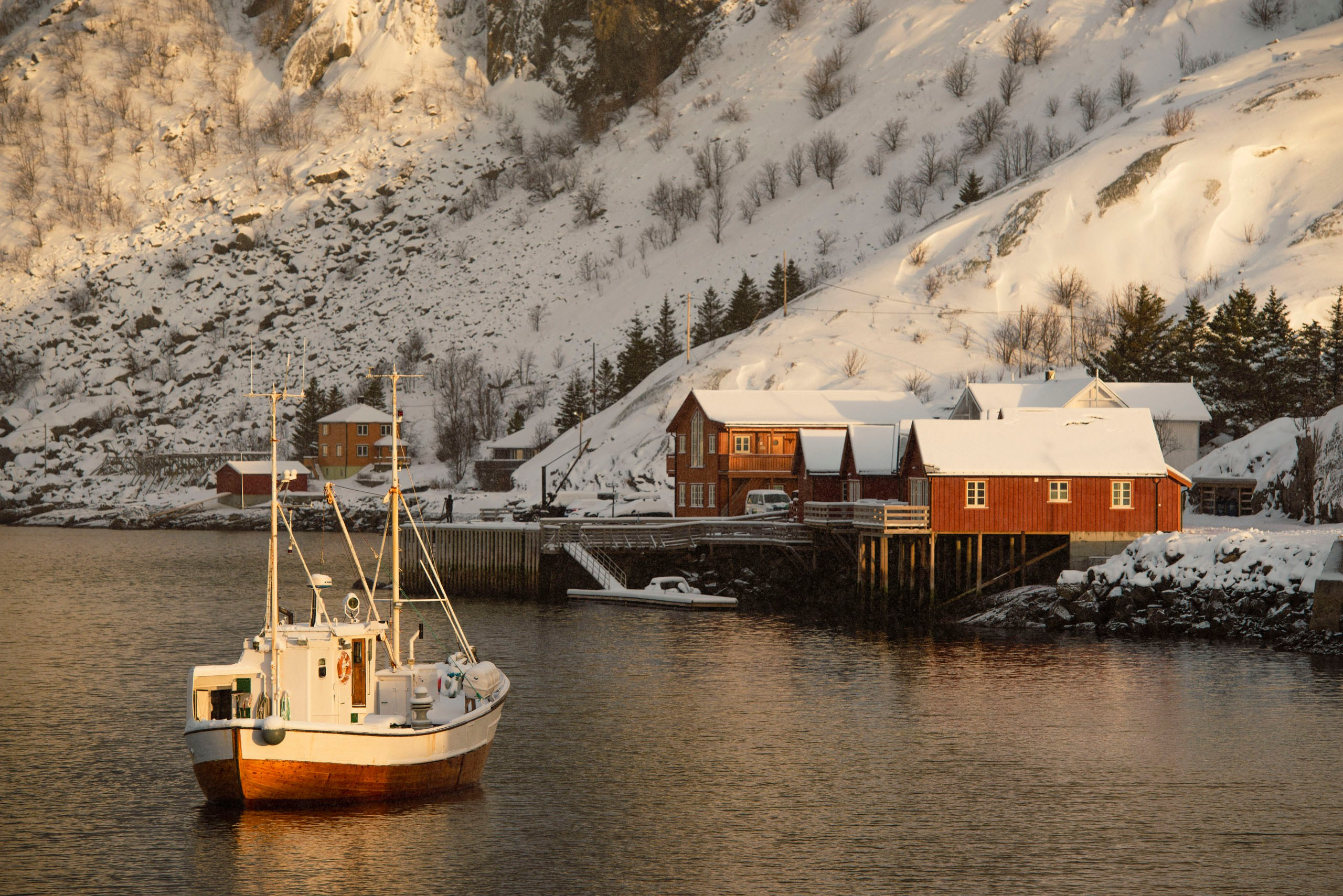 Белый рыболовецкий траулер пришвартован у деревянных пирсов красных рыбацких хижин (rorbuer) на фоне заснеженных гор в норвежской деревне Баллстад (Ballstad), Лофотенские острова.