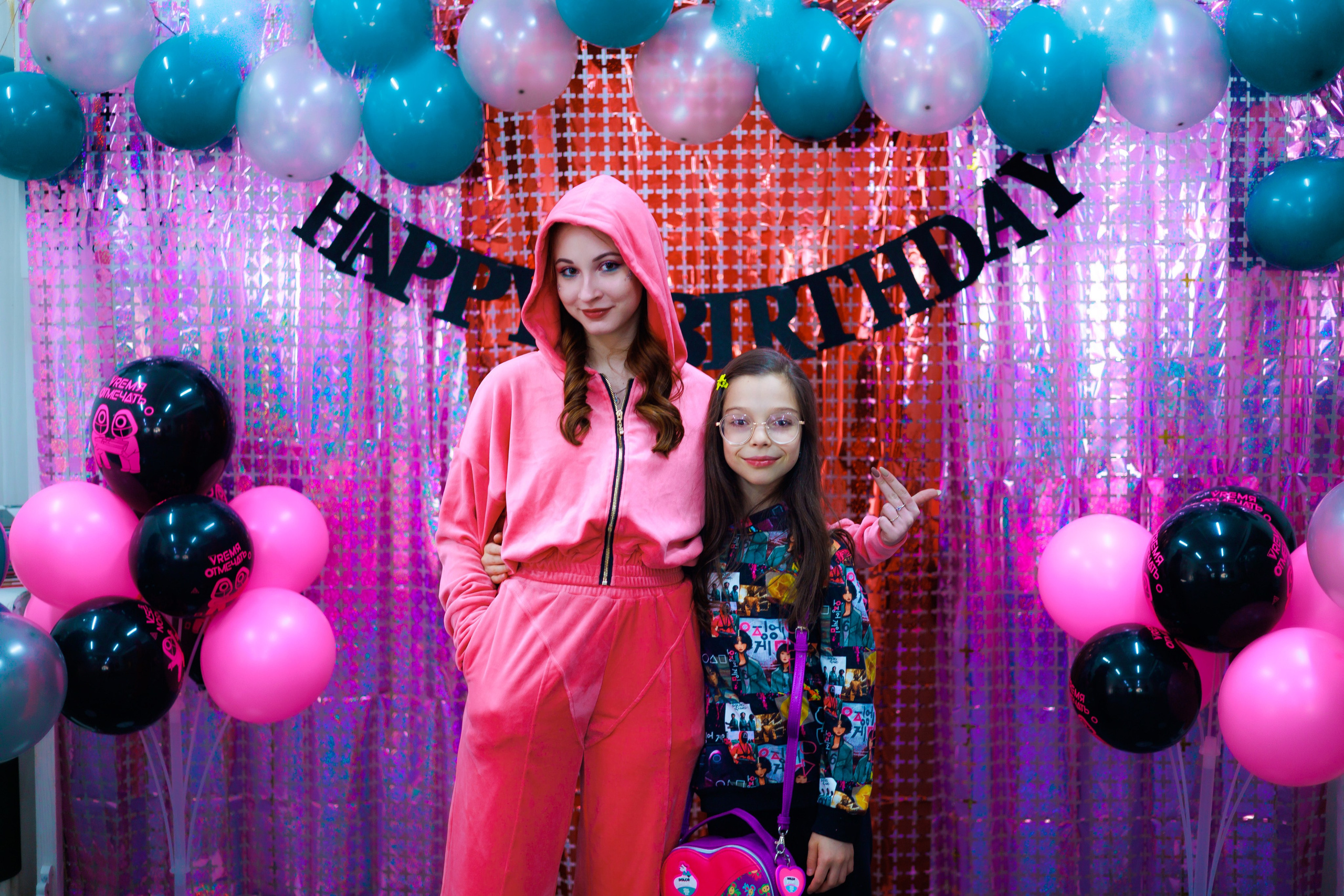 Фотозона оформлена в розовых, красных, зеленых и черных тонах специально для мероприятия в стиле Игры в кальмары. Девушка, изображающая солдата, в розовом комбинезоне позирует на фоне тематической стены с девочкой-участницей игры.