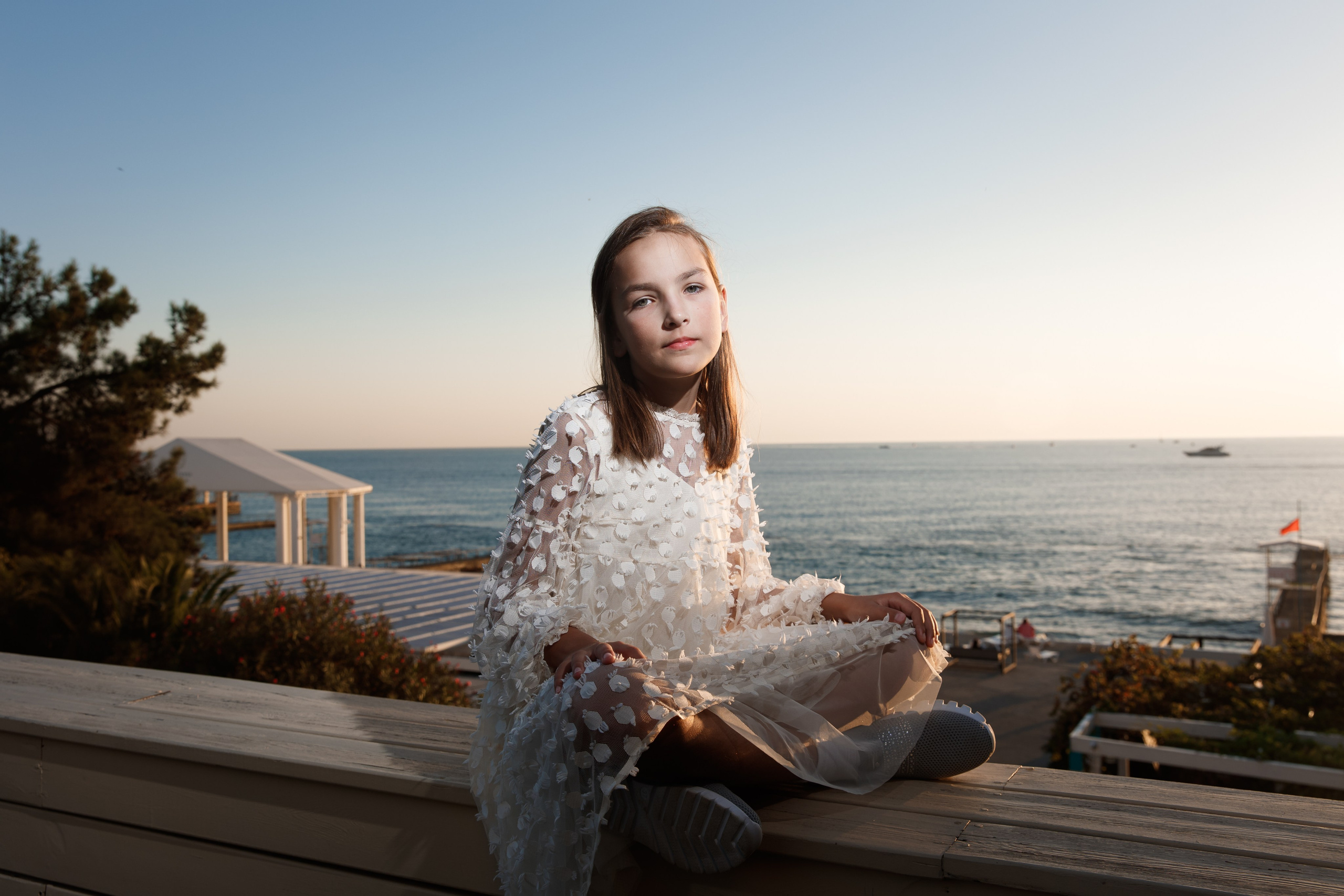 Девочка в белом платье сидит на фоне закатного моря