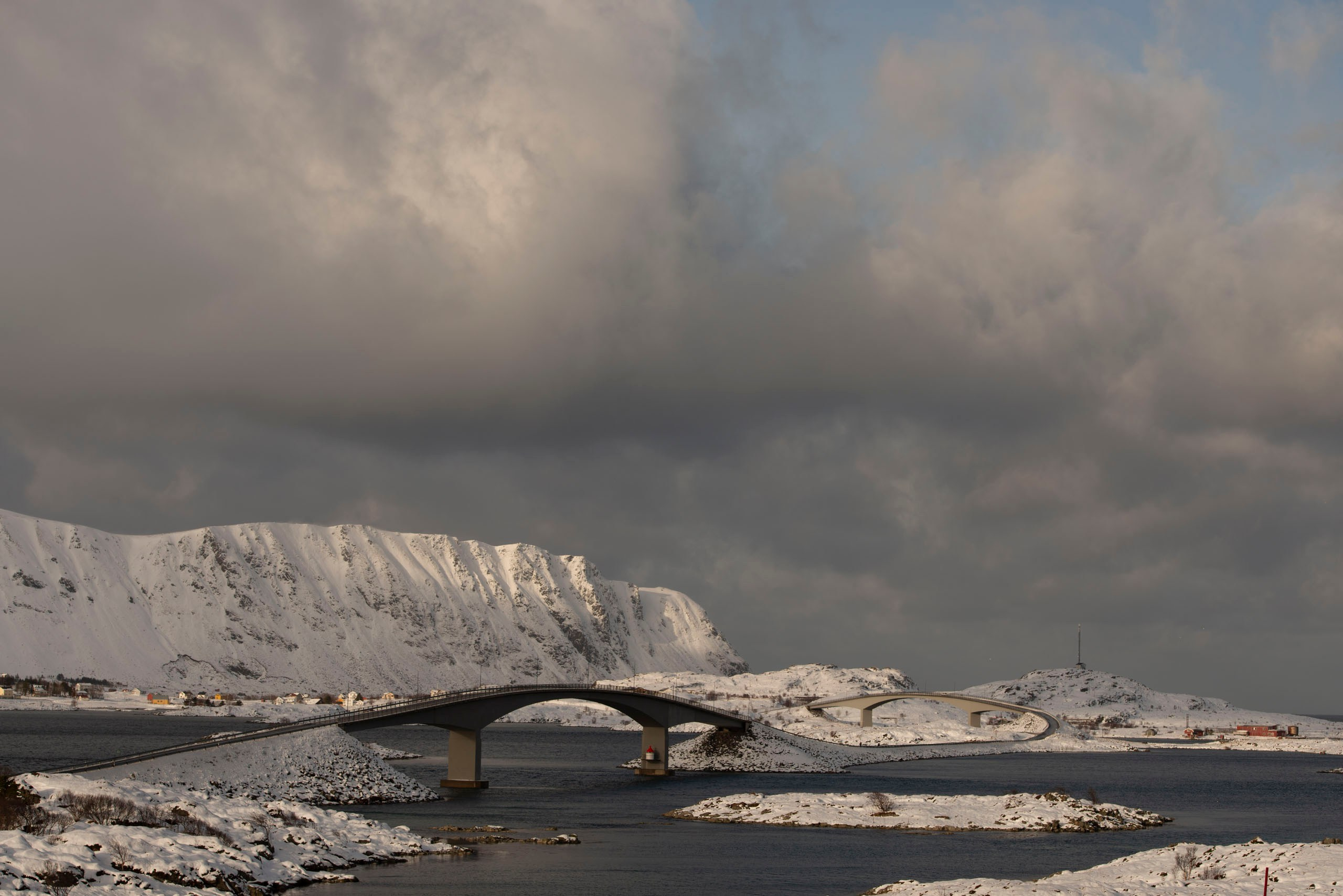 Изогнутые мосты Фредванга (Fredvang Bridges) соединяют заснеженные острова Лофотен в Норвегии на фоне гор и облачного неба.