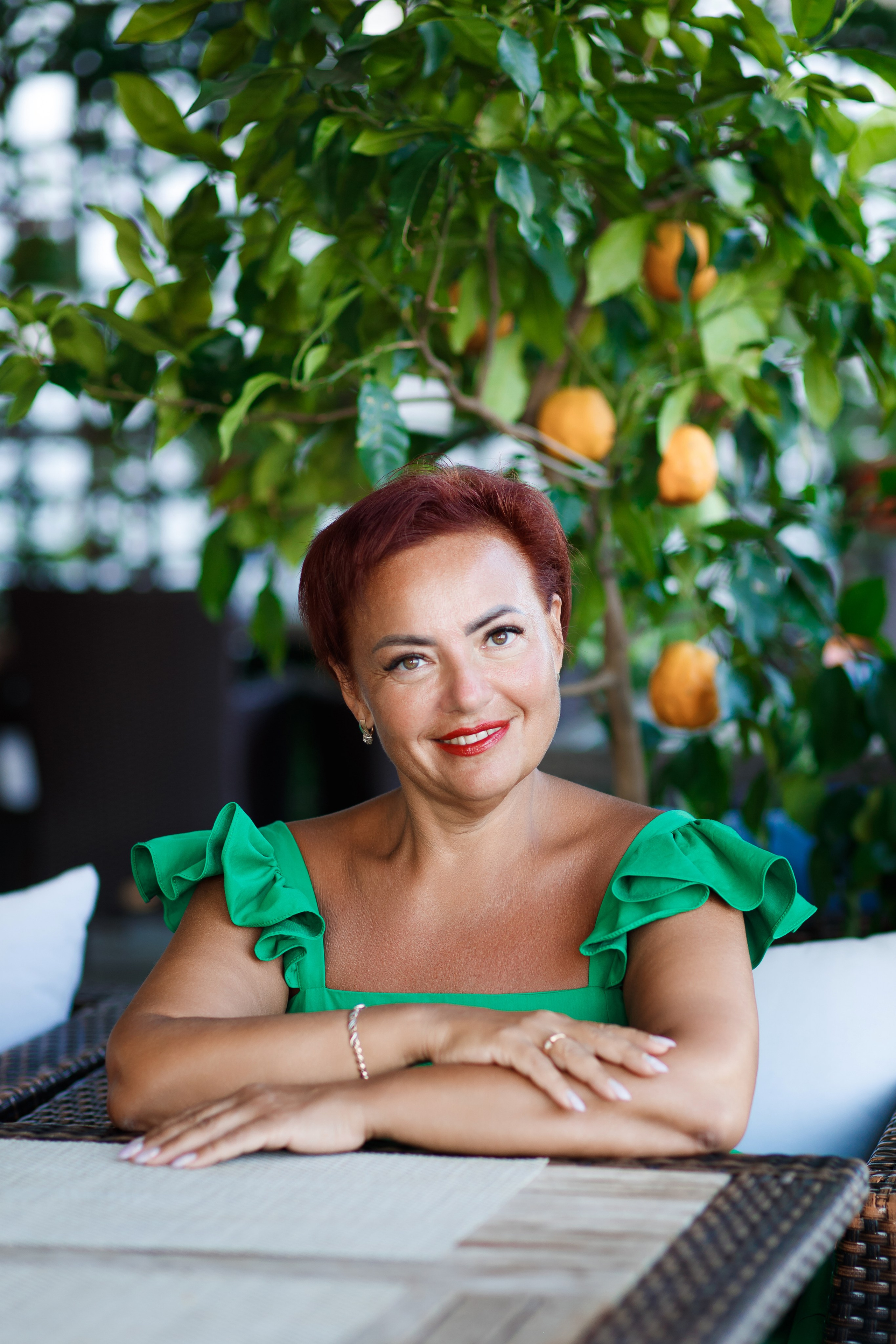 Рыжая женщина сидит за столом и улыбается смотря в камеру на фоне дерево с мандаринами
