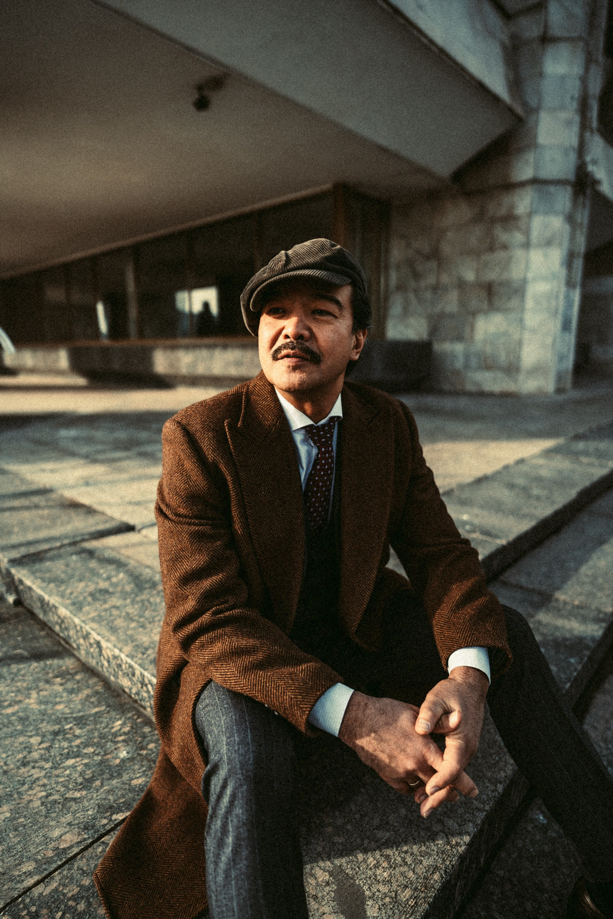 Мужчина в длинном коричневом пальто и кепке сидит на каменных ступенях в городской среде, выражая уверенность и спокойствие