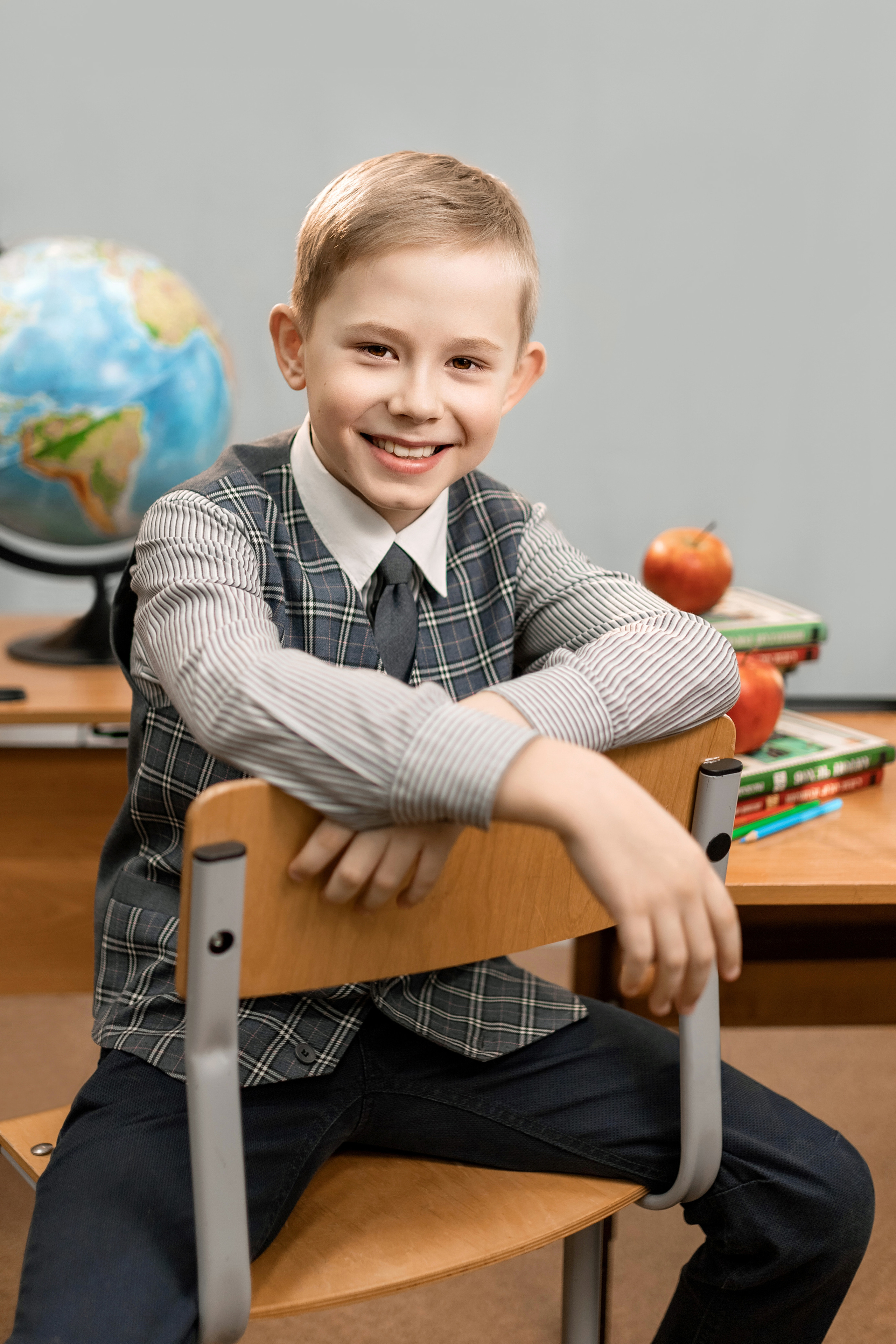 Школьник в школе сидит на стуле, а на парте лежат учебники яблоко и глобус