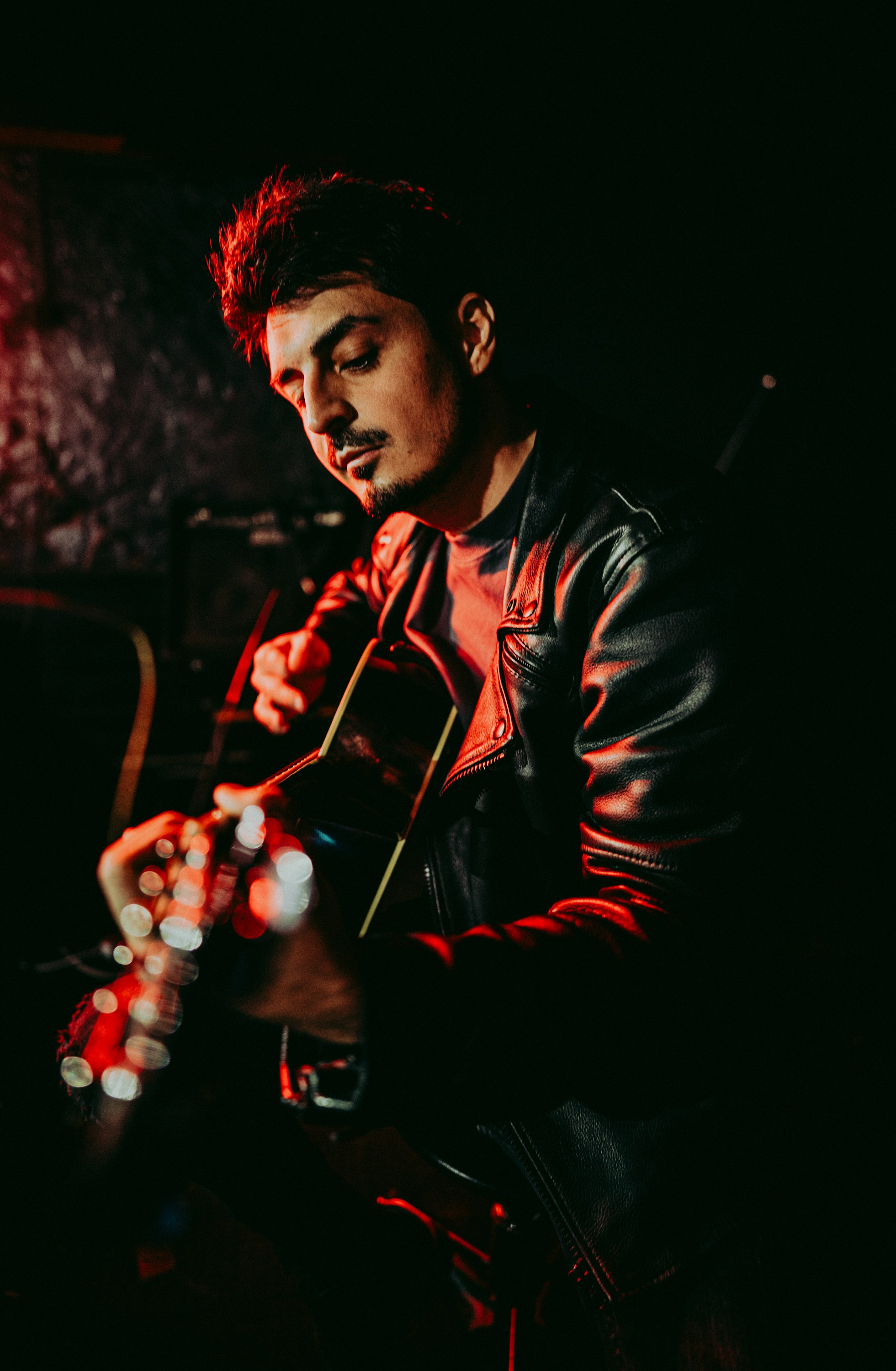Музыкант в кожаной куртке сидит в тёмной комнате и играет на гитаре, погружённый в задумчивость, с красным и чёрным светом на фоне